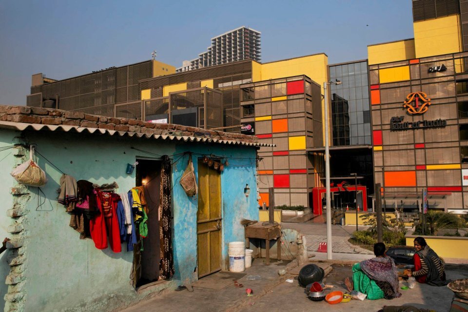 Zdjęcie przedstawia dwie kobiety ubrane w tradycyjne stroje hinduskie, siedzące na ziemi, obok porozrzucanych naczyń, przed ubogim, niebieskim, niskim budynkiem slamsów. Budynek ma dwa otwory drzwiowe, dach pokryty jest falowaną blachą, na której, na całej długości leżą cegły. Na ścianie budynku, na sznurku wiszą kolorowe ubrania. Obok budynku stoją plastikowe wiaderka i urządzenie przypominające mały piecyk, Po drugiej stronie ulicy widzimy kolorowy, przeszklony budynek nowoczesnej galerii miejskiej, a w tle nowoczesny wieżowiec.  