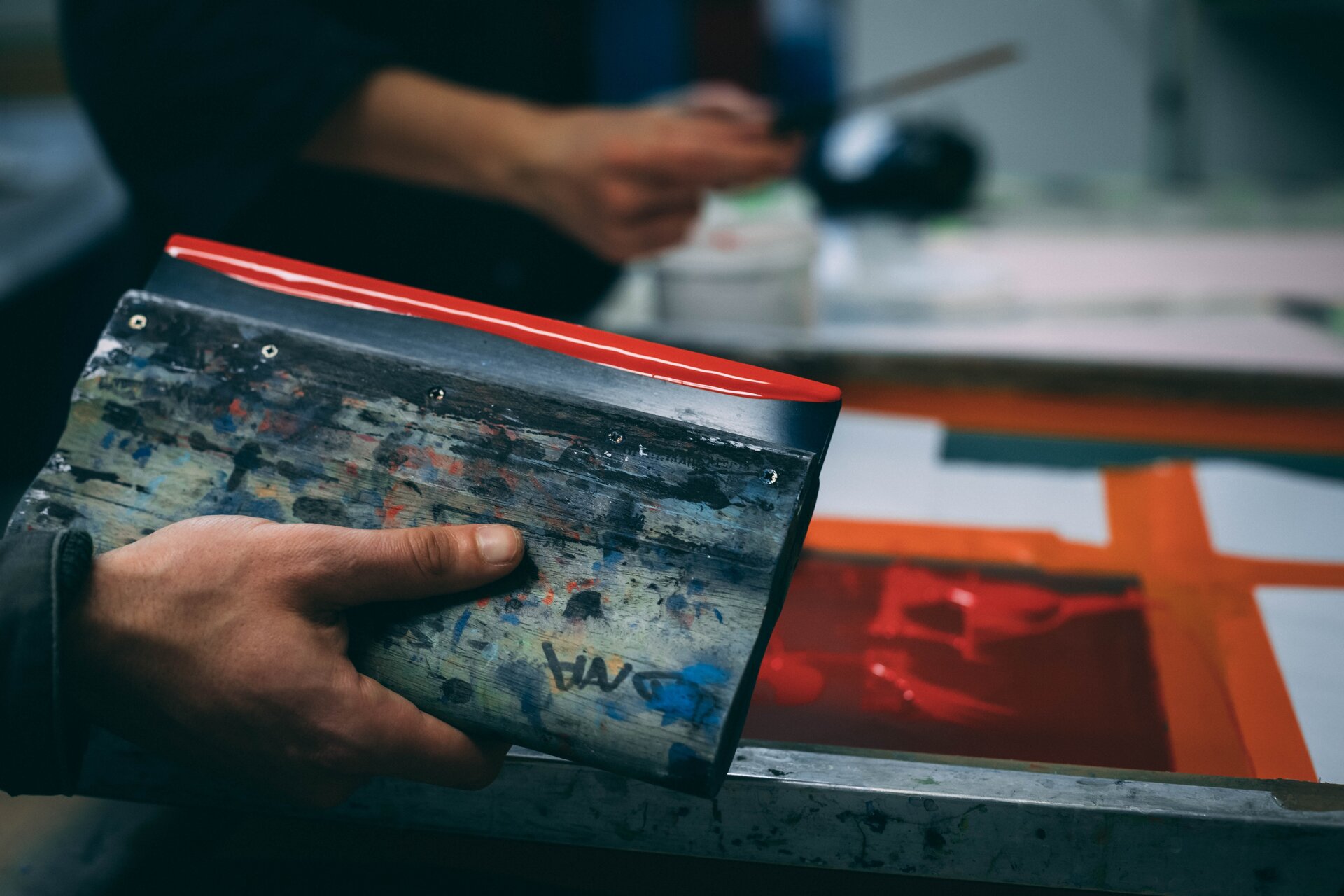 Zdjęcie przedstawia technikę sitodruku. W dolnej lewej części widoczna jest prawa dłoń z prostokątnym metalowym przedmiotem (raklem), którego dłuższa krawędź jest czerwona. W tle, powyżej, widoczna rozmazana ręka z długim przedmiotem. Po prawej widoczna forma drukowa, czyli szablon nałożony na siatkę napiętą na ramie, na który nakładana jest farba za pomocą rakla. 