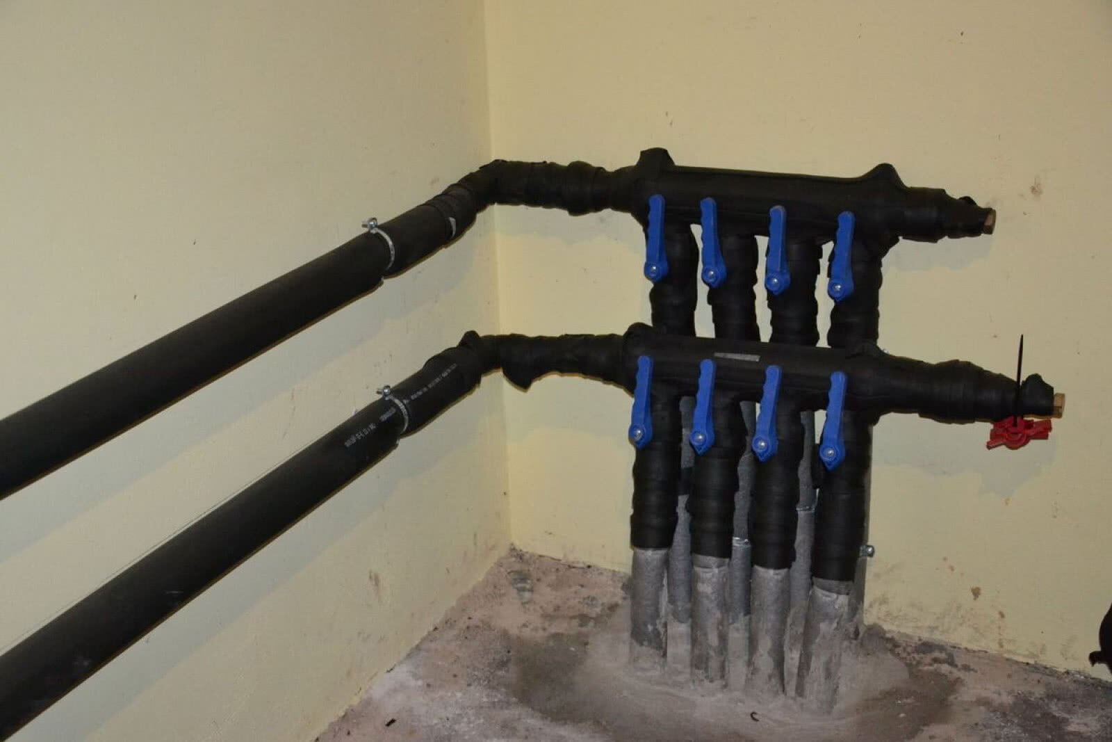 Zdjęcie przedstawia przyłącze pompy ciepła. Przy ścianie z posadzki wychodzą rury zabezpieczone otuliną. Na każdym przewodzie znajduje się zawór. Rury te zostają wprowadzone do dwóch poziomych przewodów rozmieszczonych równolegle na ścianie.