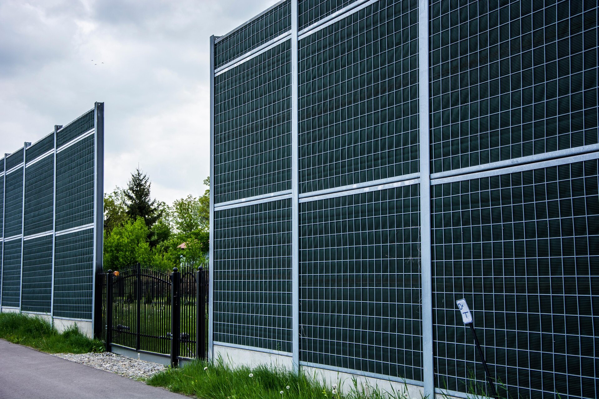 Zdjęcie przedstawia nieskuteczne zastosowanie ekranu chroniącego budynki mieszkalne przed hałasem komunikacyjnym. Widać przerwę w ciągłości ekranu. W przerwie znajduje się brama do posesji.