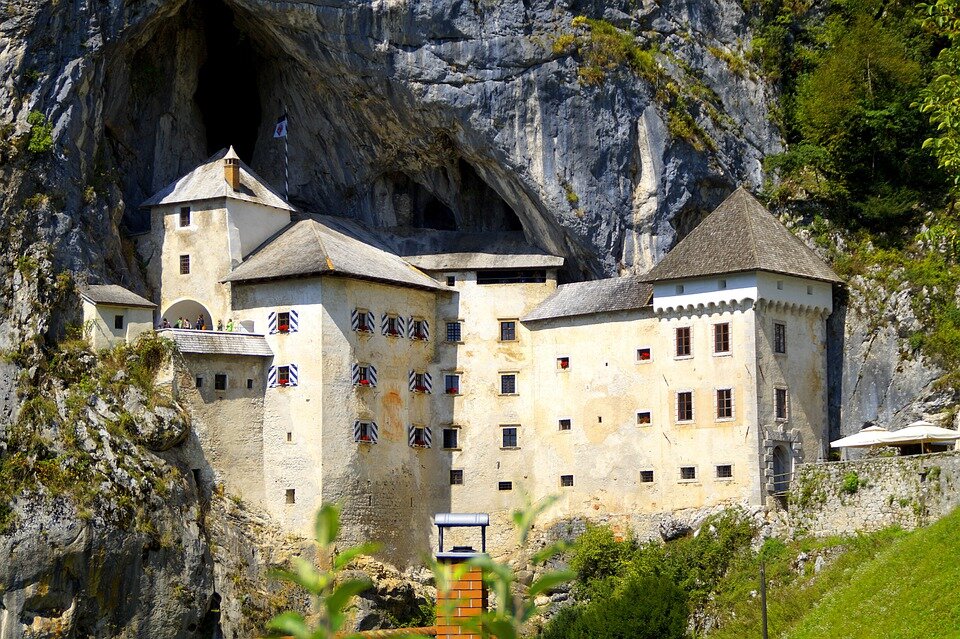 Zdjęcie przedstawia zamek usytuowany w górach skalistych delikatnie porośniętych zieloną roślinnością. W centralnej i dominującej części kadru widać zbudowany na planie litery L jasnobeżowy zamek z dwoma wieżyczkami. Zamek ma brązowy spiczasty dach i niewielkie okna. W jego lewej części okna mają okiennice w biało‑granatowe pasy. Zdjęcie jest jasne, wykonane w pogodny dzień.