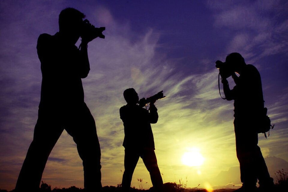 Zdjęcie przedstawia trzy sylwetki fotografujących mężczyzn na tle słońca. Każdy stoi odwrócony w inną stronę, jakby wyczekując odpowiedniej chwili na zrobienie zdjęcia. Patrzą przez swoje aparaty fotograficzne.