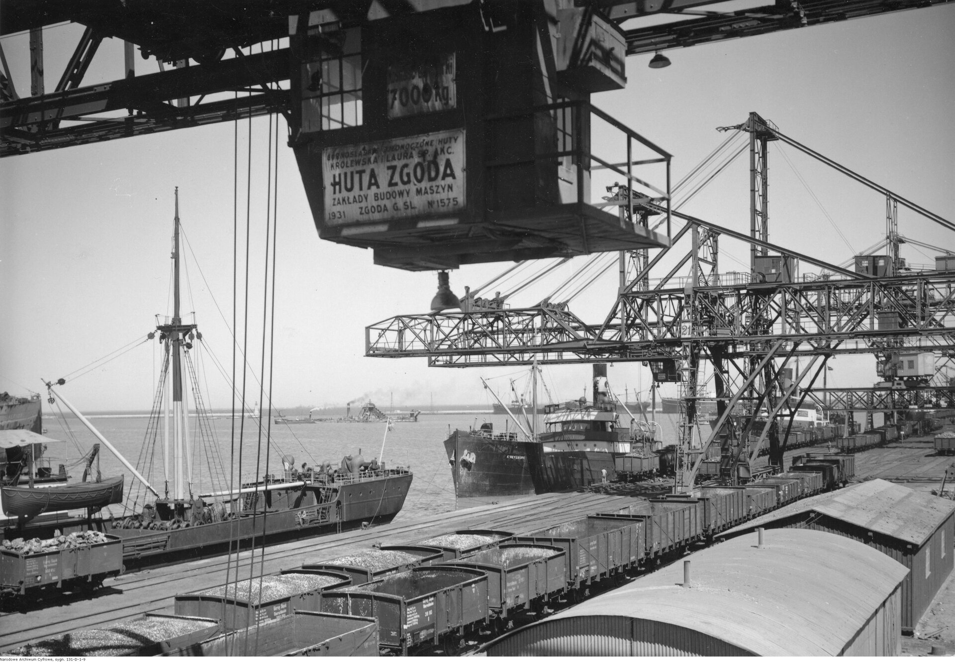 Zdjęcie przedstawia port z molem transportowym, na którym stoją otarte wagony towarowe, w których znajduje się węgiel. Przy rampie stoją zacumowane statki towarowe, nad wszystkim widoczne dźwigi. Na pierwszym planie element dźwigu z napisem: Huta Zgoda.