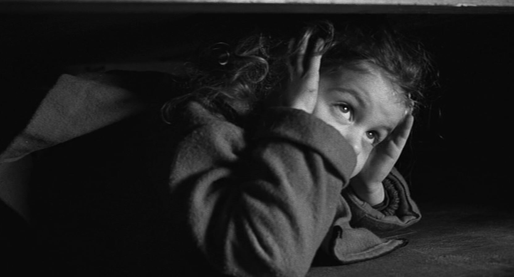 Zdjęcie przedstawia leżącą na podłodze, na brzuchu, dziewczynkę. Dziecko zasłania uszy dłońmi. Patrzy się w górę.