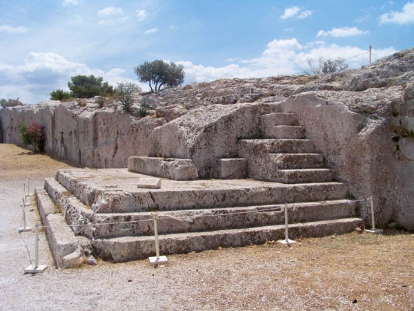 Zdjęcie przedstawia ruiny starożytnej budowli. Na pierwszym planie widoczne są schody i zniszczony fragment podestu, na którym ustawiona była mównica.