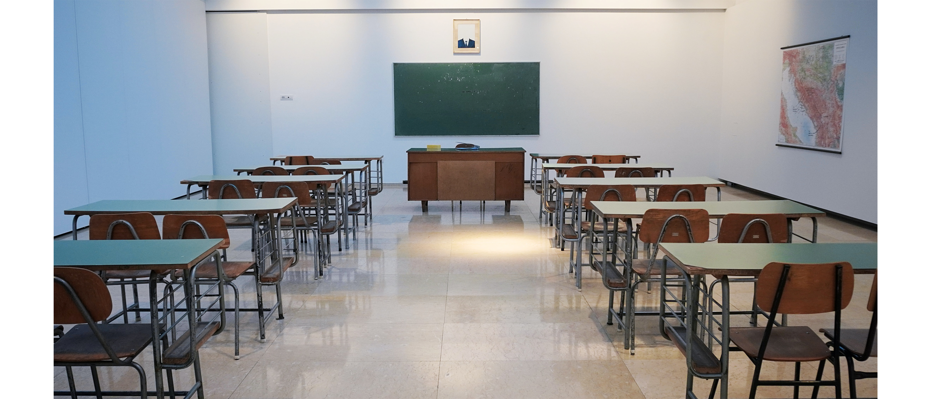 Zdjęcie przedstawia pustą salę klasową w szkole. Po lewej i po prawej stronie stoją szkolne ławki i krzesła. W centralnej części wisi tablica, przed którą znajduje się biurko.