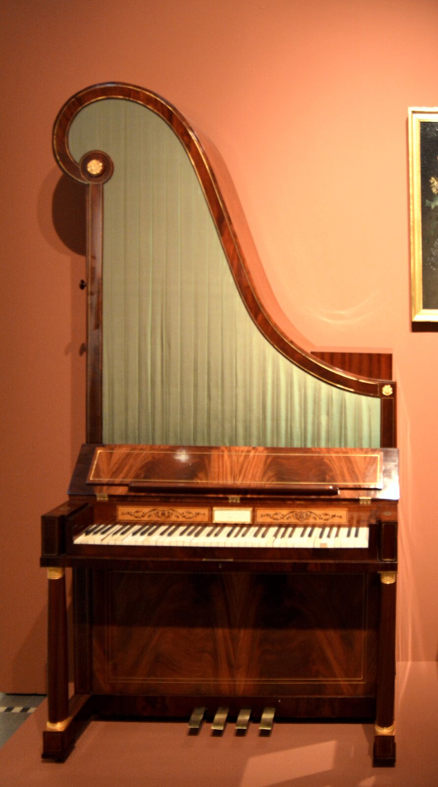 Zdjęcie przedstawia drewniany instrument przypominający pianino, ale z ustawionym pionowo wysokim, zaokrąglonym elementem w kształcie trójkąta, przypominającym harfę. Instrument posiada białe i czarne klawisze oraz cztery pedały. 