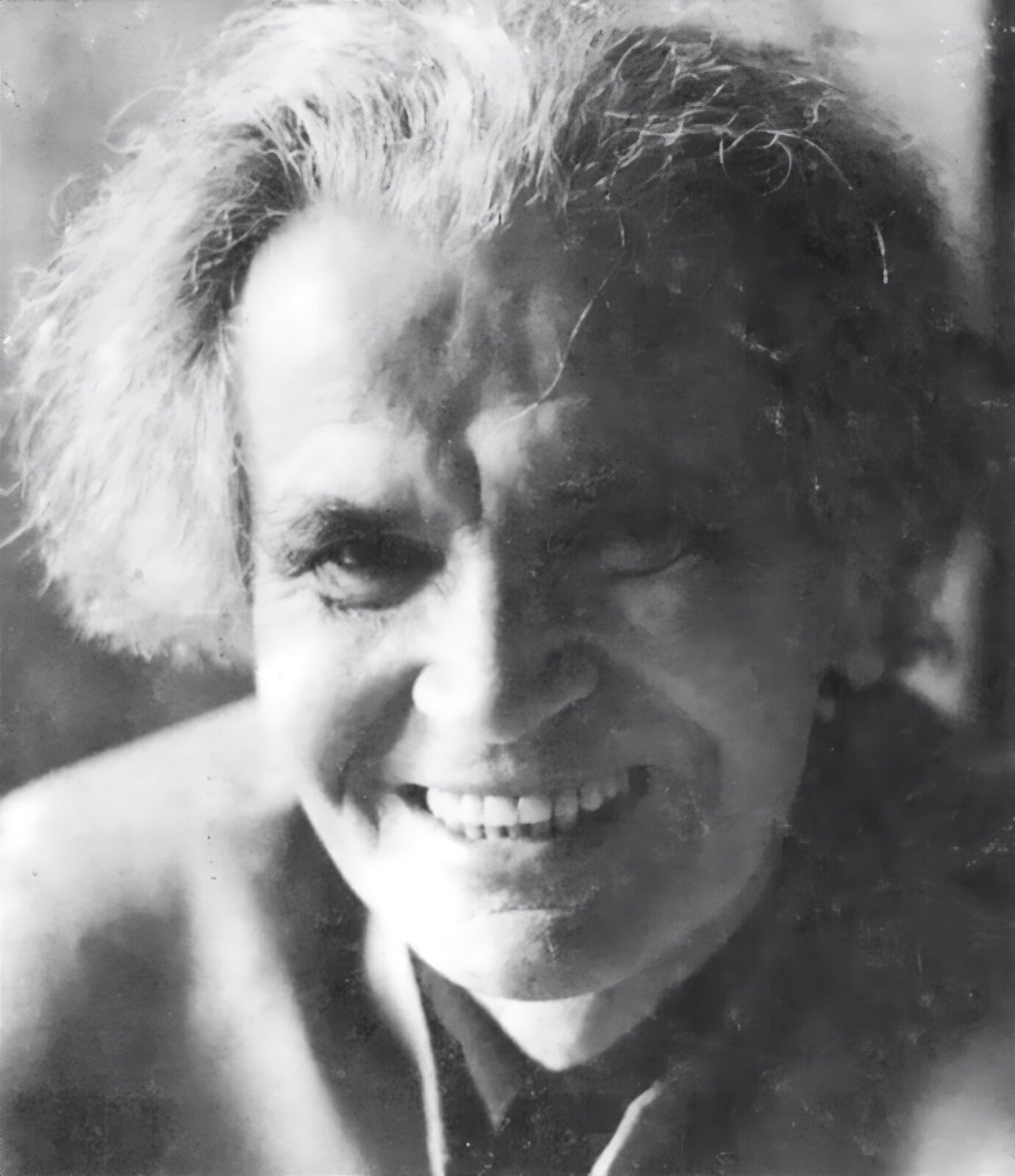 Czarno‑biały portret starszego mężczyzny o średniej długości siwych, zaczesanych do tyłu włosach, ciemnych oczach i szerokim uśmiechu.