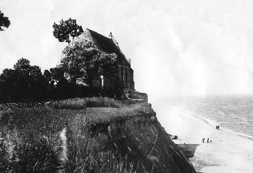 Archiwalne, czarno-białe zdjęcie kościoła w Trzęsaczu z 1870 r. Na nadmorskim urwisku stoi prostopadłościenna budowla z dwuspadowym, spiczastym dachem. Strome urwisko łączy się u podnóża z plażą. W tle morze. 