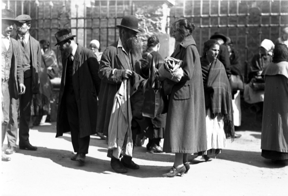 Kliknij aby powiększyć Na zdjęciu scena handlu ulicznego. Na środkowym planie widać mężczyznę z długą, gęstą brodą, ubranego w kapelusz, koszulę, kamizelkę i długi płaszcz. Trzyma on w prawej ręce jasną oraz ciemną marynarkę. Obok mężczyzny stoi kobieta dorównująca mu wzrostem. Ubrana jest w długi płaszcz oraz czółenka na niskim obcasie. W lewej ręce kobieta trzyma zwinięte w rulon ubrania i czarną, skórzaną torbę. Na drugim planie widoczni są przechadzający się po ulicy mężczyźni i kobiety.