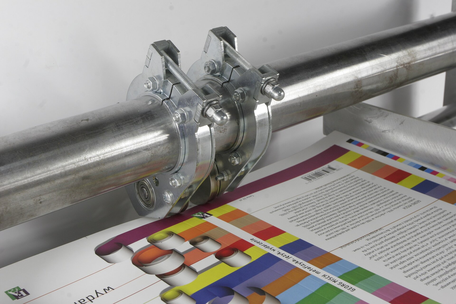 Zdjęcie przedstawia przykładowy półautomat do bigowania. Na pierwszym planie widoczne jest zadrukowane podłoże, ułożone na stole. W tle widoczna jest stalowa rura biegnąca w poziomie, na której znajdują się dwa pierścienie, które dotykają podłoża drukowego.