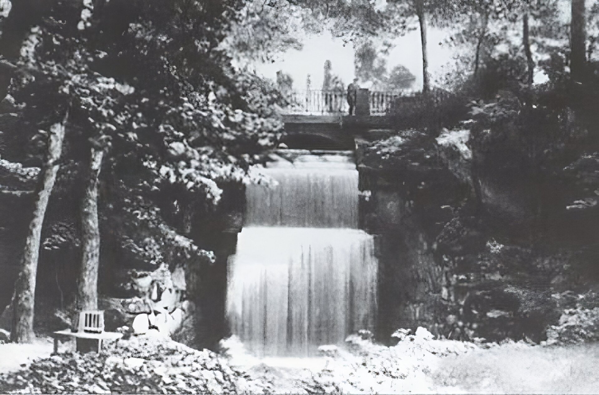 Kliknij, aby powiększyć. Zdjęcie przedstawia widok kaskadowego wodospadu na terenie parku „Zofiówka” w Humaniu z mostkiem widokowym nad nim. Po obu stronach wodospadu widoczne są aleje parkowe z ławeczkami dla kontemplacji pięknych widoków. Wokół roztacza się bujna roślinność parkowa.