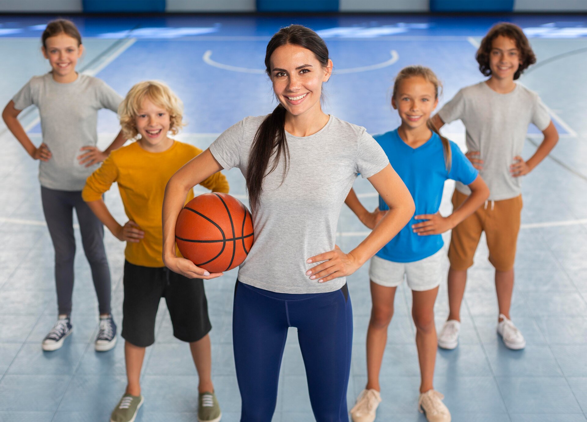 Zdjęcie przedstawia kobietę w młodym wieku stojącą na sali gimnastycznej. Trzyma ona pod pachą piłkę do gry w koszykówkę. Za nią widoczna jest czwórka dzieci w wieku szkolnym. 
