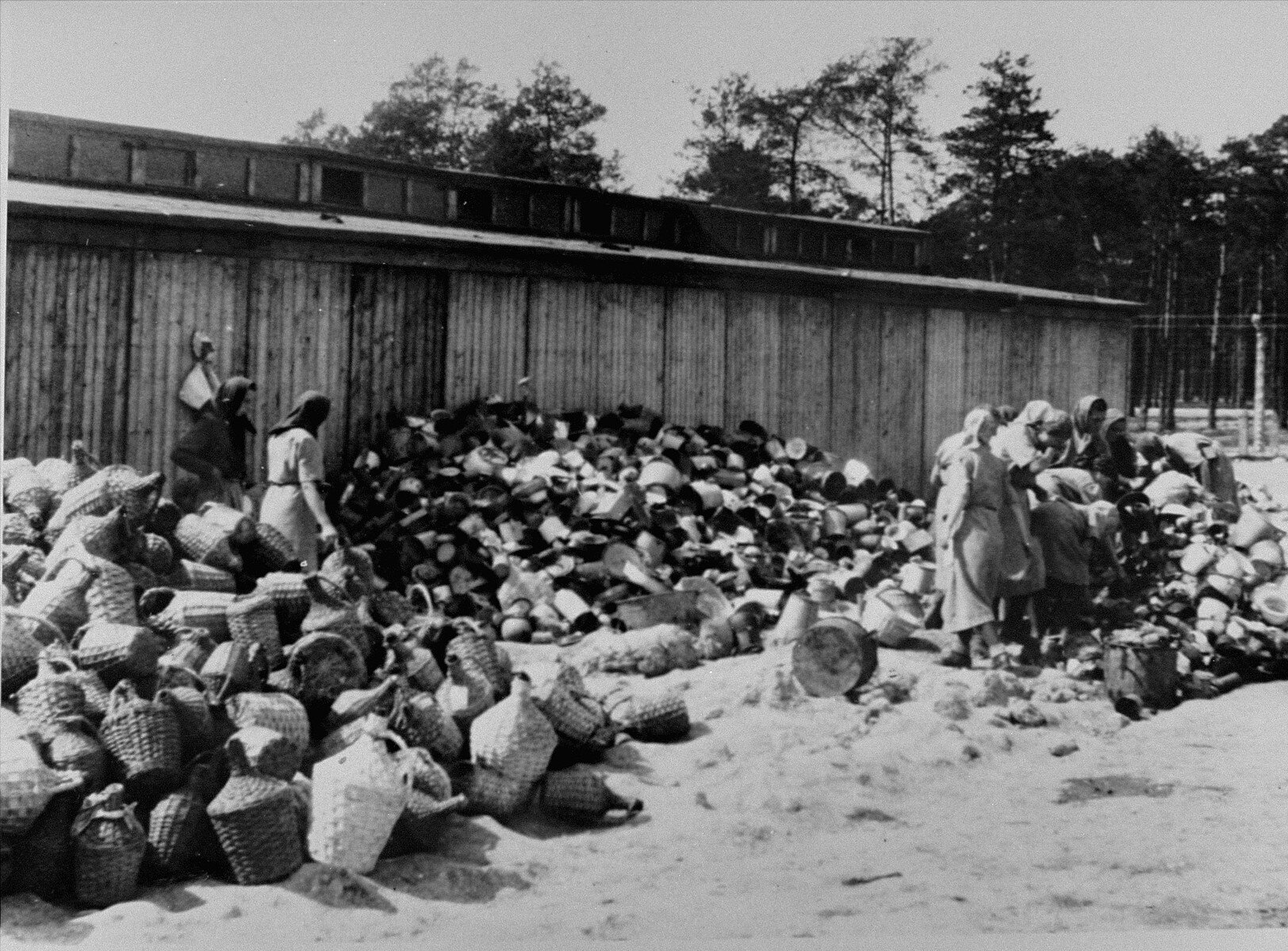 Zdjęcie przedstawia fragment placu przed obozowym barakiem. Na placu są zgromadzone różne rzeczy w stosach: balony w wiklinowych koszach, wiadra, kosze. Przedmioty sortują znajdujące się na placu kobiety.