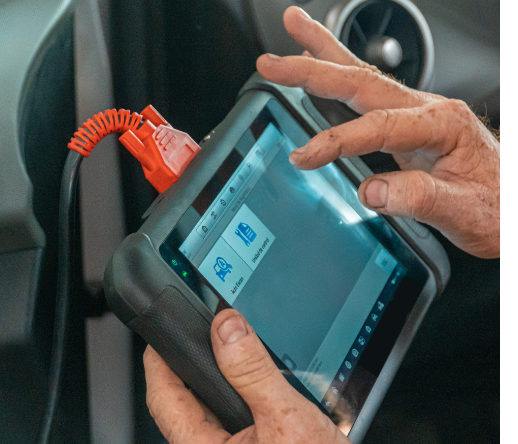 Na zdjęciu ukazana dłoń obsługująca komputerowy system diagnostyczny, czyli urządzenie podobne do tabletu podpięte kablem do pojazdu.