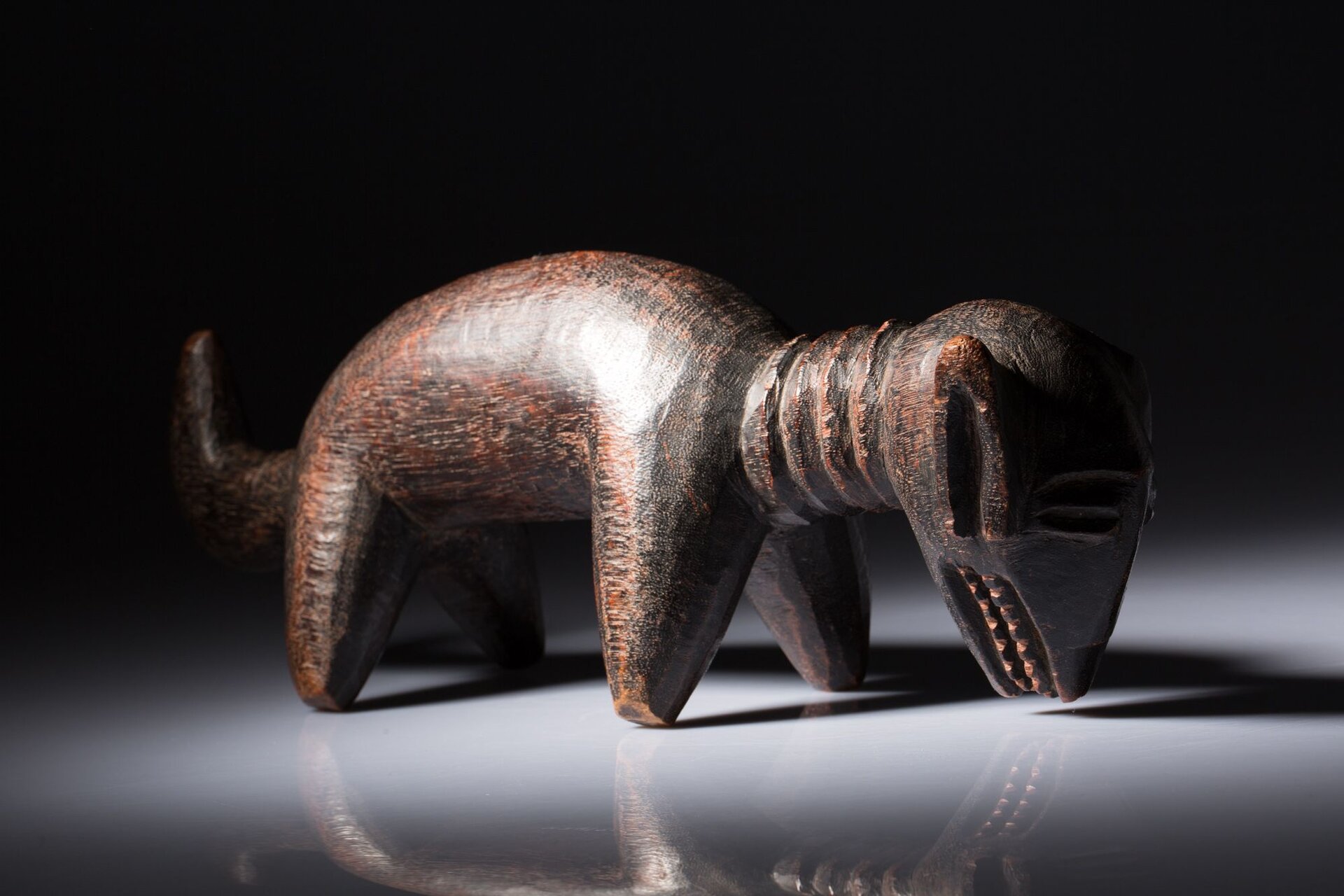 Zdjęcie przedstawia rzeźbę psa. Jest ona wykonana z drewna w kolorze ciemnobrązowym. Przedstawia zwierzę stojące bokiem. Po lewej stronie jest ogon, a po prawej głowa. Ogon ma podniesiony do góry. Zwierzę stoi na czterech nogach- szerokich na górze i wąskich na nodze. Ma długą szyję z zaznaczonymi 4 okręgami. Głowę ma podłużną, opuszczoną w dół. Zaznaczone są długie uszy, oczy i zęby. Rzeźba stoi na szarym, błyszczącym blacie na tle czarnej ściany. Na blacie jest cień i odbicie rzeźby. 