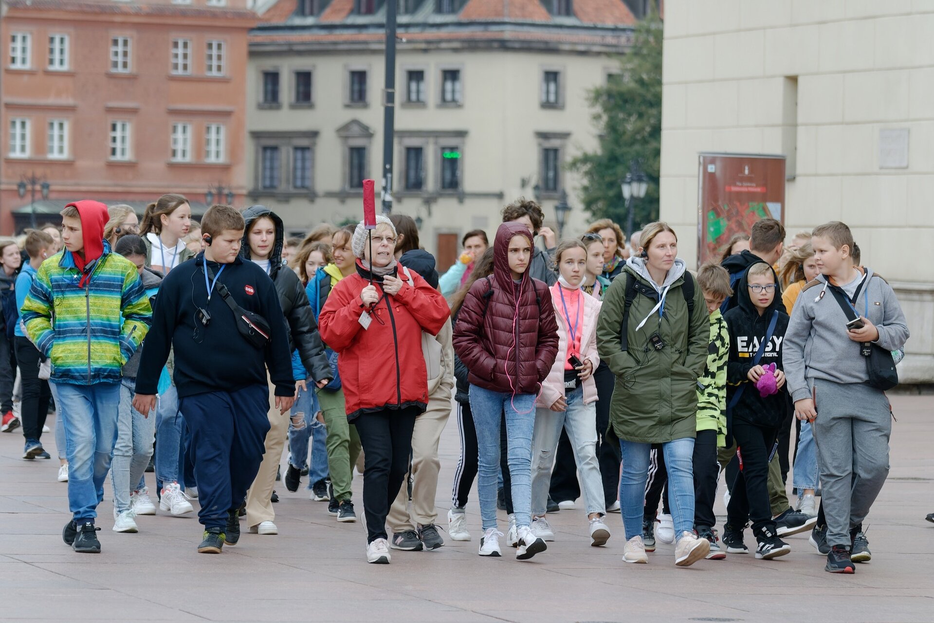 Ilustracja przedstawia grupę młodych turystów spacerujących po mieście. Przewodniczka idzie  z przodu, trzymając w dłoni czerwoną parasolkę.