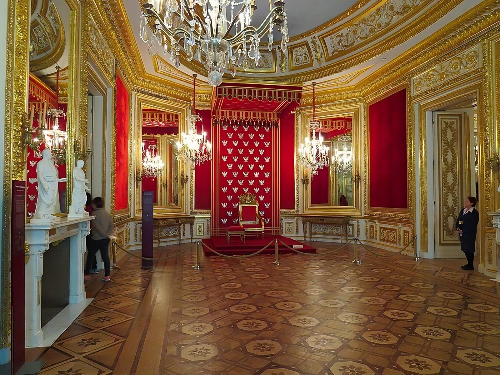 Kliknij, aby powiększyć Zdjęcie przedstawia pomieszczenie audiencyjne króla z wyeksponowanym tronem z podnóżkiem, kory znajduje się przy środkowej ścianie. Na ścianie za tronem wisi zaplecek tronu, zdobiony haftowanymi srebrem na tle z czerwonego aksamitu. Wokół tronu są wielkie lustra w złoconych ramach. Przy lewej ścianie stoi marmurowy kominek, zdobiony antycznymi rzeźbami. W prawej ścianie umieszczone są drzwi. Są otwarte i wchodzi przez nie człowiek.