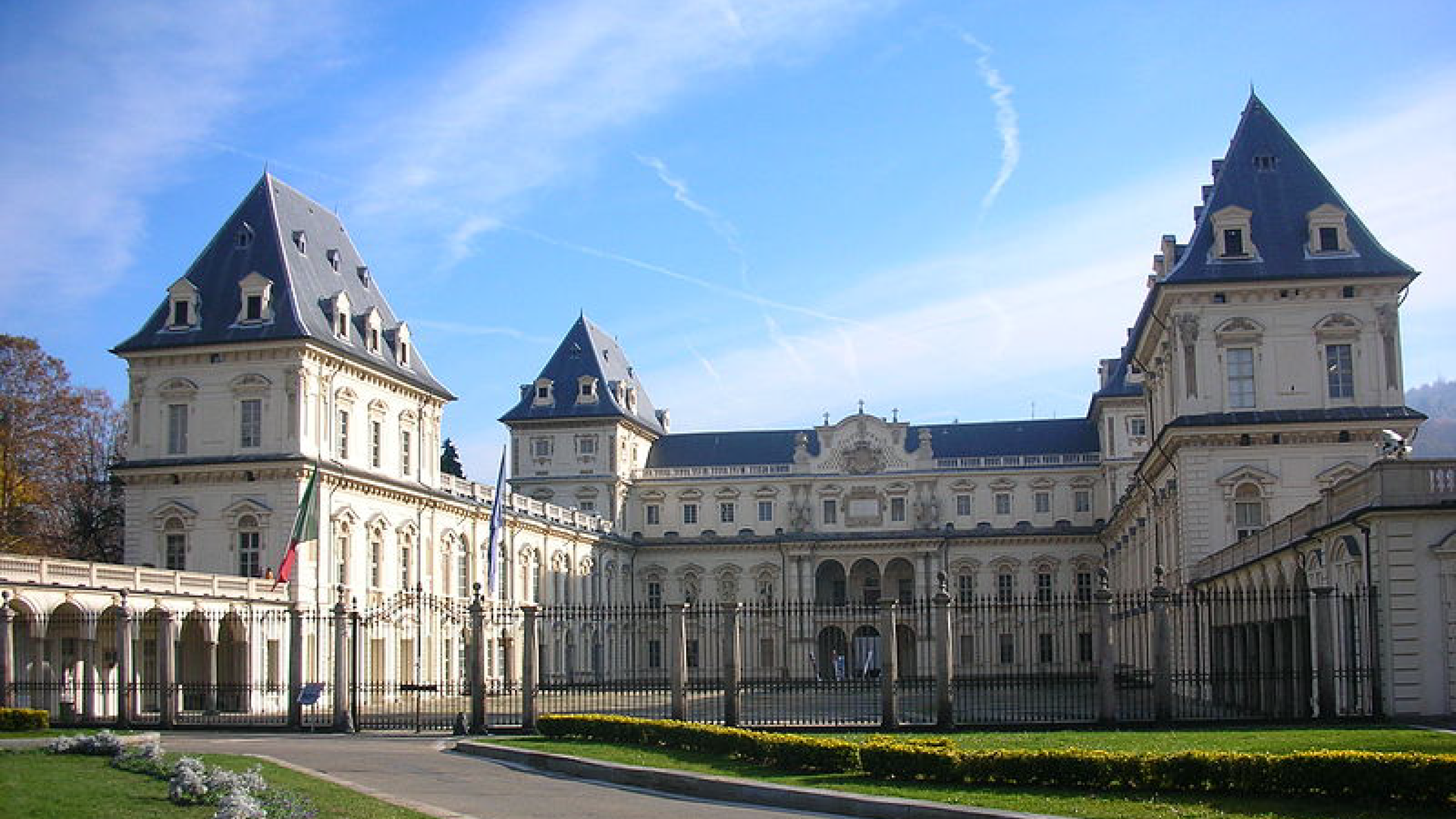Zdjęcie przedstawia budynek Politechniki Turyńskiej. Budynek jest kilkupiętrowy, ma szpiczasty, opadający na dwie strony dach. Budynek jest w kształcie litery U. Przed budynkiem znajduje się dziedziniec.
