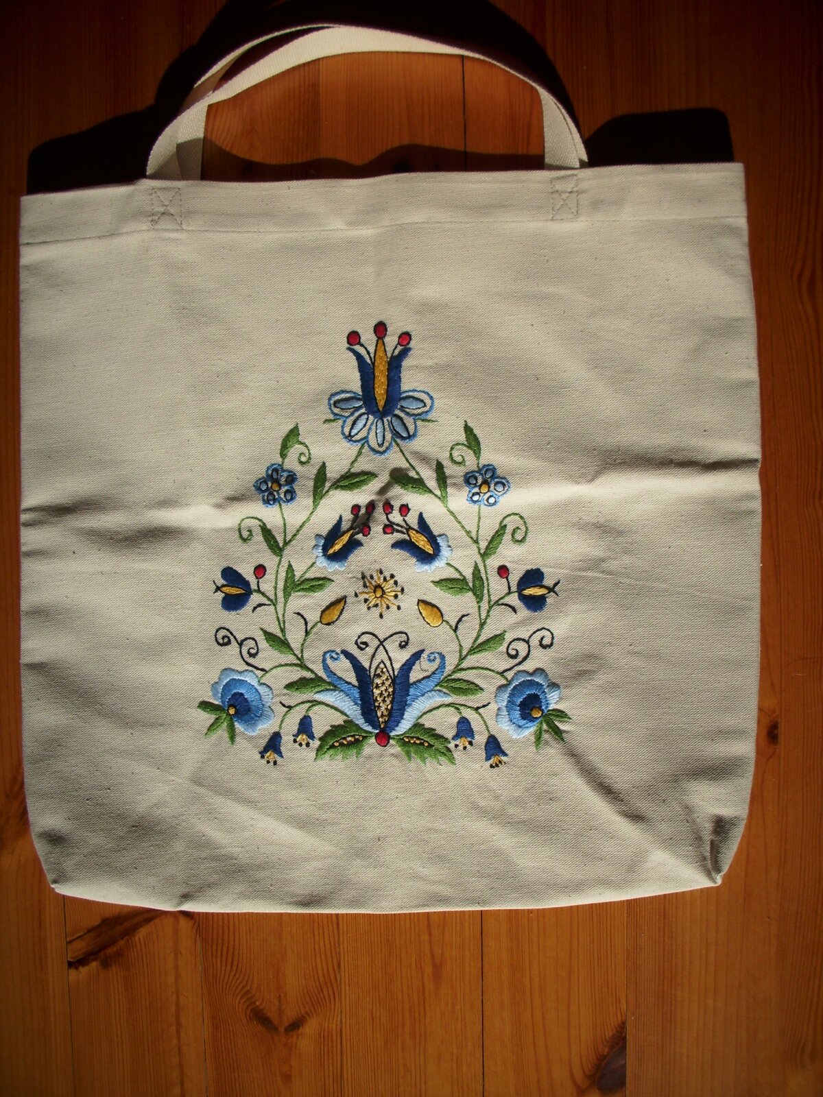 Zdjęcie przedstawia torbę wielokrotnego użytku. Jest na niej wyhaftowany wzór kaszubski - niebieskie kwiaty i gałązki z liśćmi.  