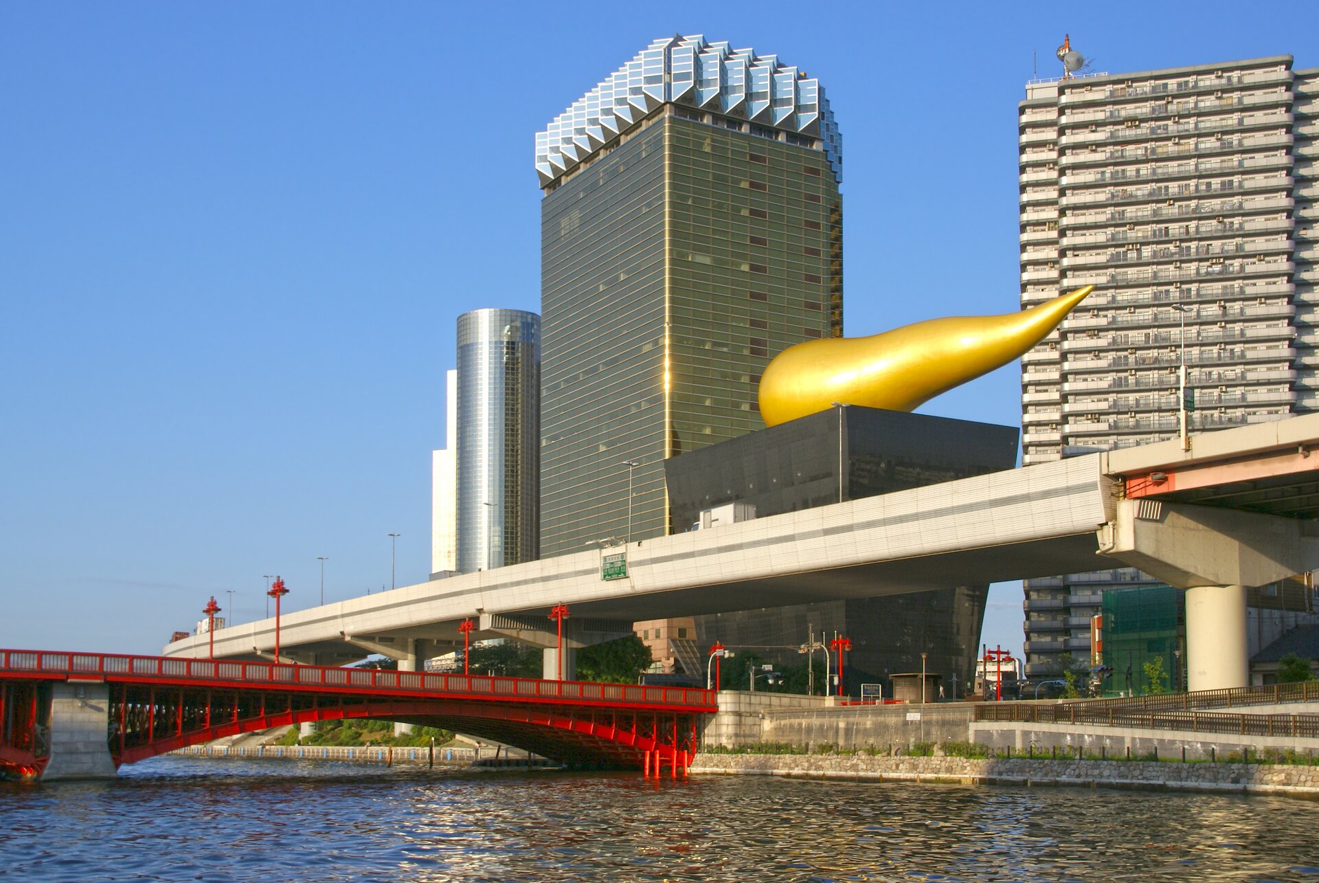 Fotografia barwna w układzie poziomym przedstawiająca fragment panoramy Tokio w słoneczny dzień z leżącym na dachu jednego z budynków w centrum kadru charakterystycznym obiektem w kształcie złotego poziomego płomienia.
Budynek rozszerza się ku górze. Stojący za nim z lewej strony trochę jaśniejszy, szary, wysoki biurowiec posiada złocone elementy narożne i zwieńczenie z jasnych, szklanych okien ustawionych w trójkątnych modułach na jego szczycie, tworzących coś w rodzaju zębatki. Prawą stronę kadru wypełnia jasny wieżowiec oddzielony od opisanego wcześniej błękitną przestrzenią nieba, które wypełnia większą część górnej części kadru. U dołu na pierwszym planie przebiega betonowa estakada. Jeszcze bliżej kanał z przerzuconym przez niego czerwonym mostem, który zaczynając się w środku kadru, niknie za lewą krawędzią fotografii.