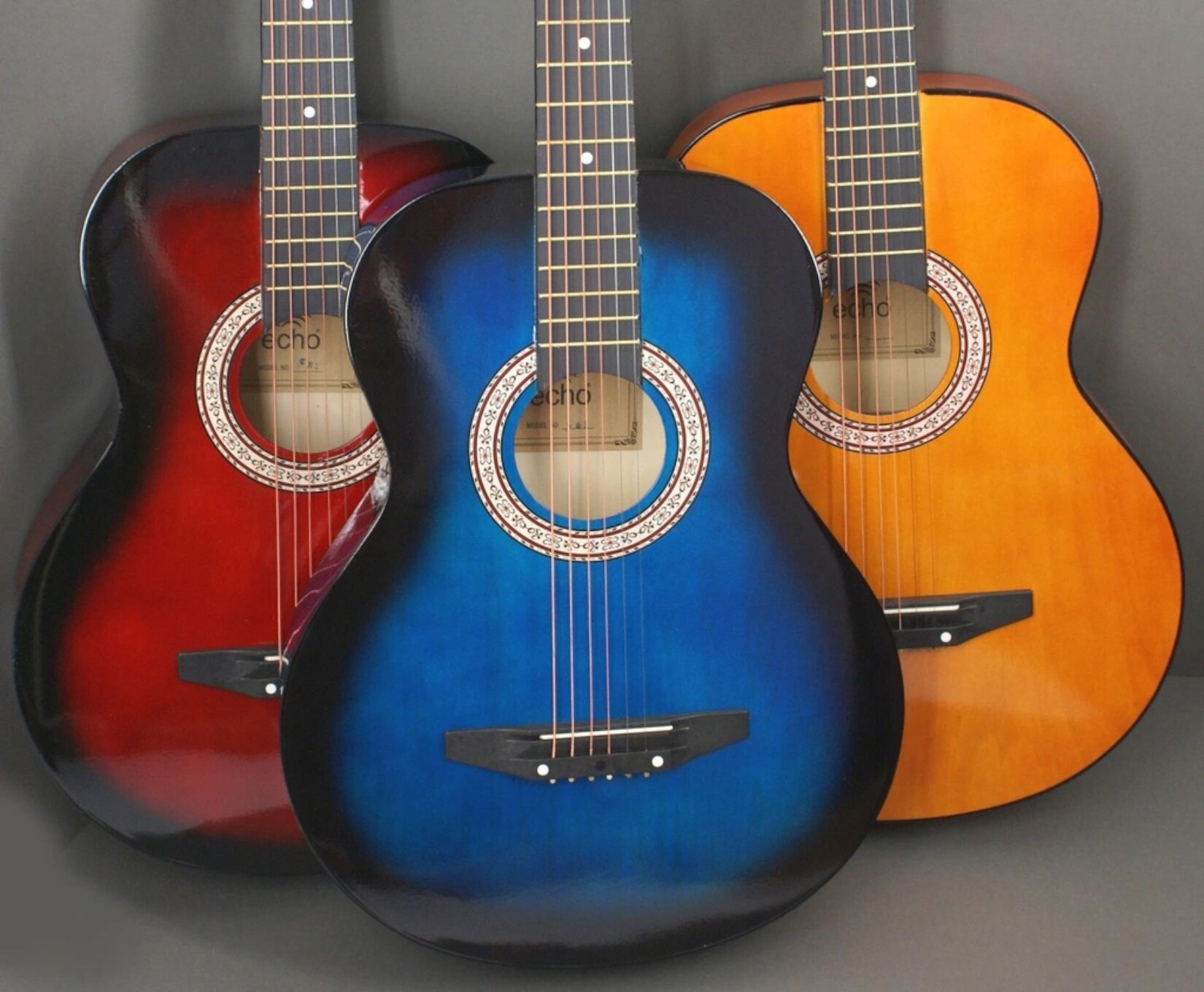 Ilustracja przedstawia trzy gitary klasyczne, stojące na szarym tle. Gitara po lewej stronie jest czerwona z czarnym podpaleniem. Gitara po środku jest niebieska z czarnym podpaleniem, a gitara po prawej stronie jest pomarańczowa.