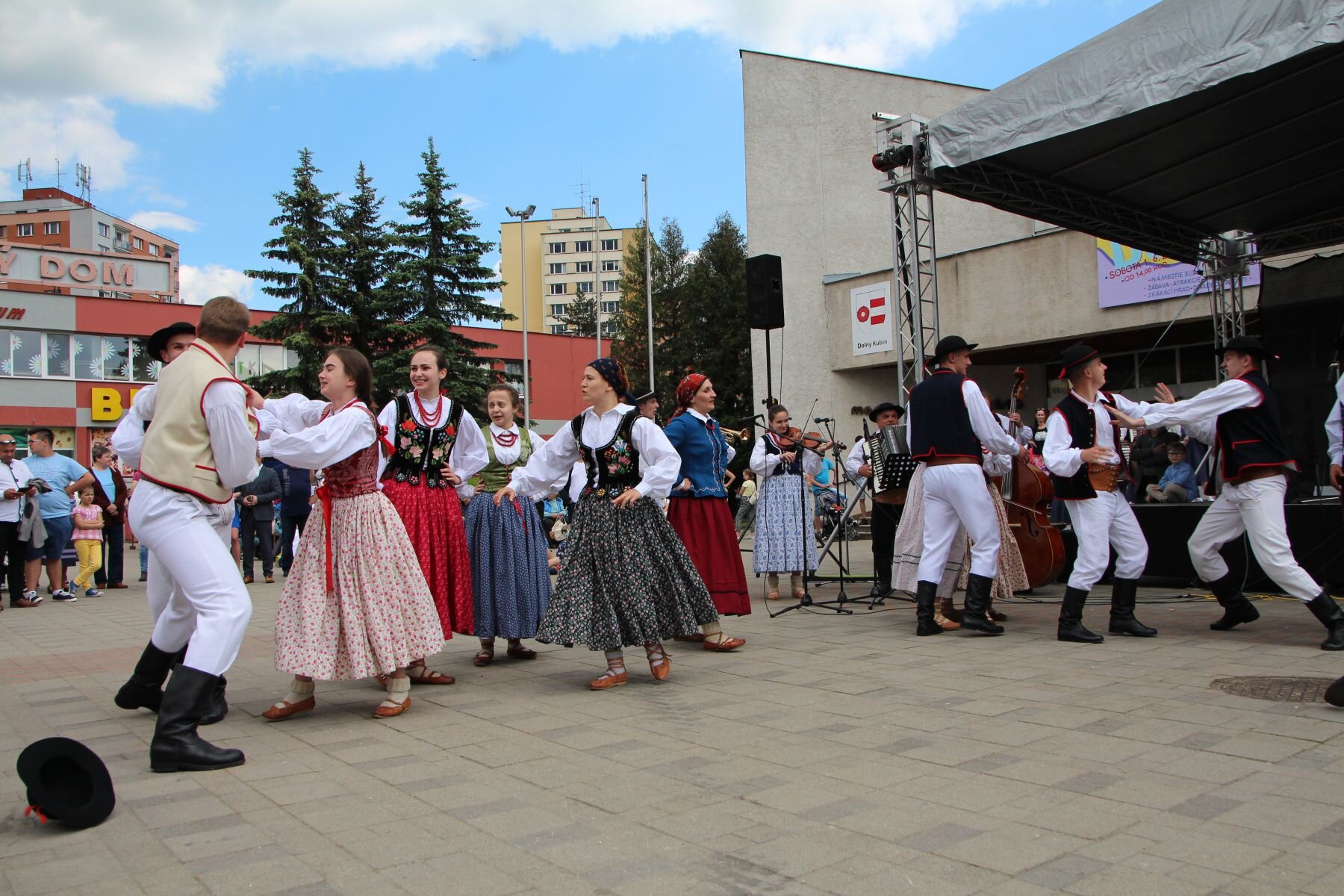 Na zdjęciu przedstawiona jest grupa osób ubranych w tradycyjne stroje limanowskie. 
Z lewej strony stoją dwaj mężczyźni oraz grupa kobiet. Jeden z mężczyzn ubrany jest w kremową kamizelkę, białe spodnie oraz czarne buty. Na ziemi, z lewej strony leży kapelusz mężczyzny. Kobiety ubrane są w różnokolorowe suknie oraz fartuszki - białe, czerwone, niebieskie, czarne. Stroje zdobione są kwiecistymi wzorami. Na szyjach kobiet wiszą czerwone korale. Z prawej strony, pod sceną - mężczyźni. Ubrani są w czarne buty, białe spodnie, białe koszule i czarne kamizelki z czerwonym dekorem oraz czarne kapelusze. Mężczyźni stoją blisko siebie, wyprostowani. Jeden z mężczyzn wyciąga ręce do drugiego. Tło stanowi scena oraz budynki. Na scenie można dostrzec muzyków grających na skrzypcach, harmonii i kontrabasie. 