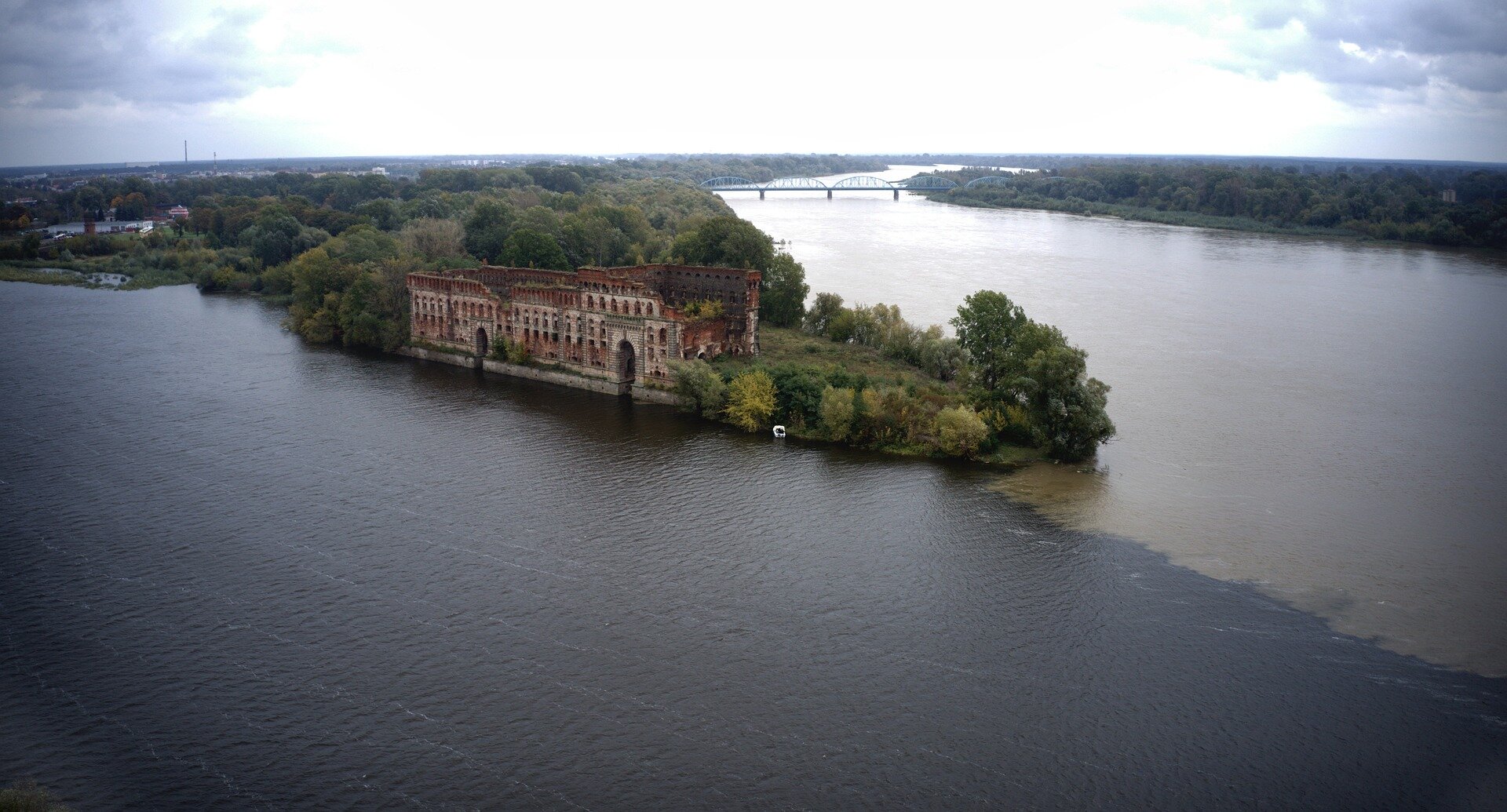 Fotografia przedstawia ujście Narwi do Wisły. Narew wpada do Wisły po prawej stronie. Wisłę od Narwi oddziela trójkątny półwysep porośnięty drzewami, na którym zlokalizowany jest fragment ceglanych zabudowań twierdzy.