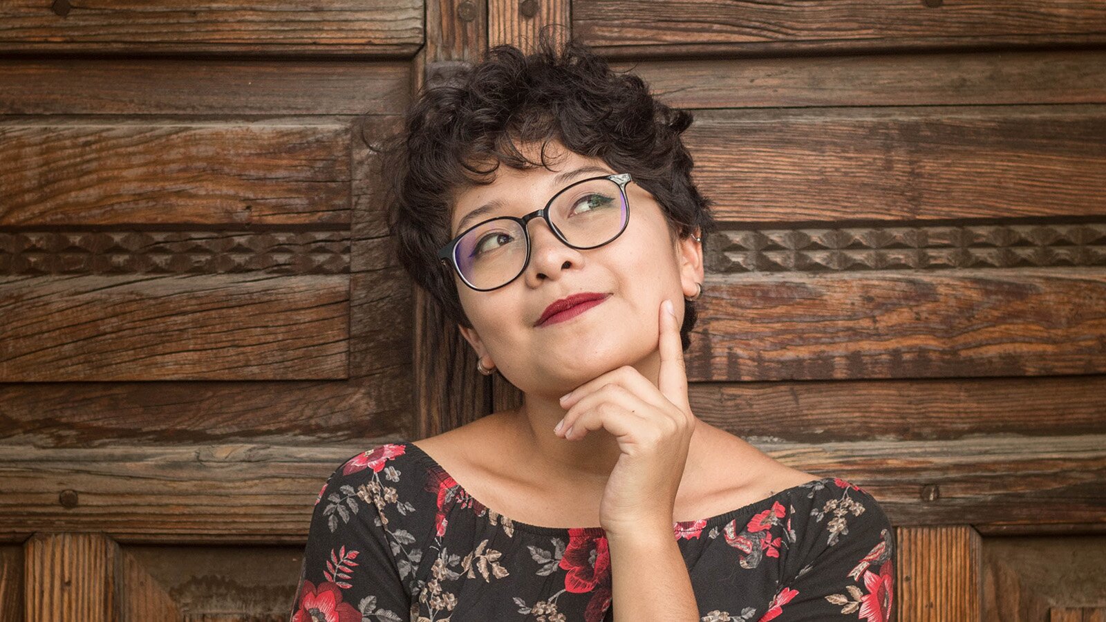 Zdjęcie przedstawia popiersie kobiety w średnim wieku. Głowa kobiety oparta jest o drewnianą ścianę i jest lekko uniesiona ku górze. Widoczna jest prawa dłoń, która dotyka brody, a palec wskazujący opiera się na żuchwie. Wzrok kobiety jest rozmarzony, skierowany w prawo górną stronę. Pomalowane na czerwono usta są w lekkim uśmiechu.