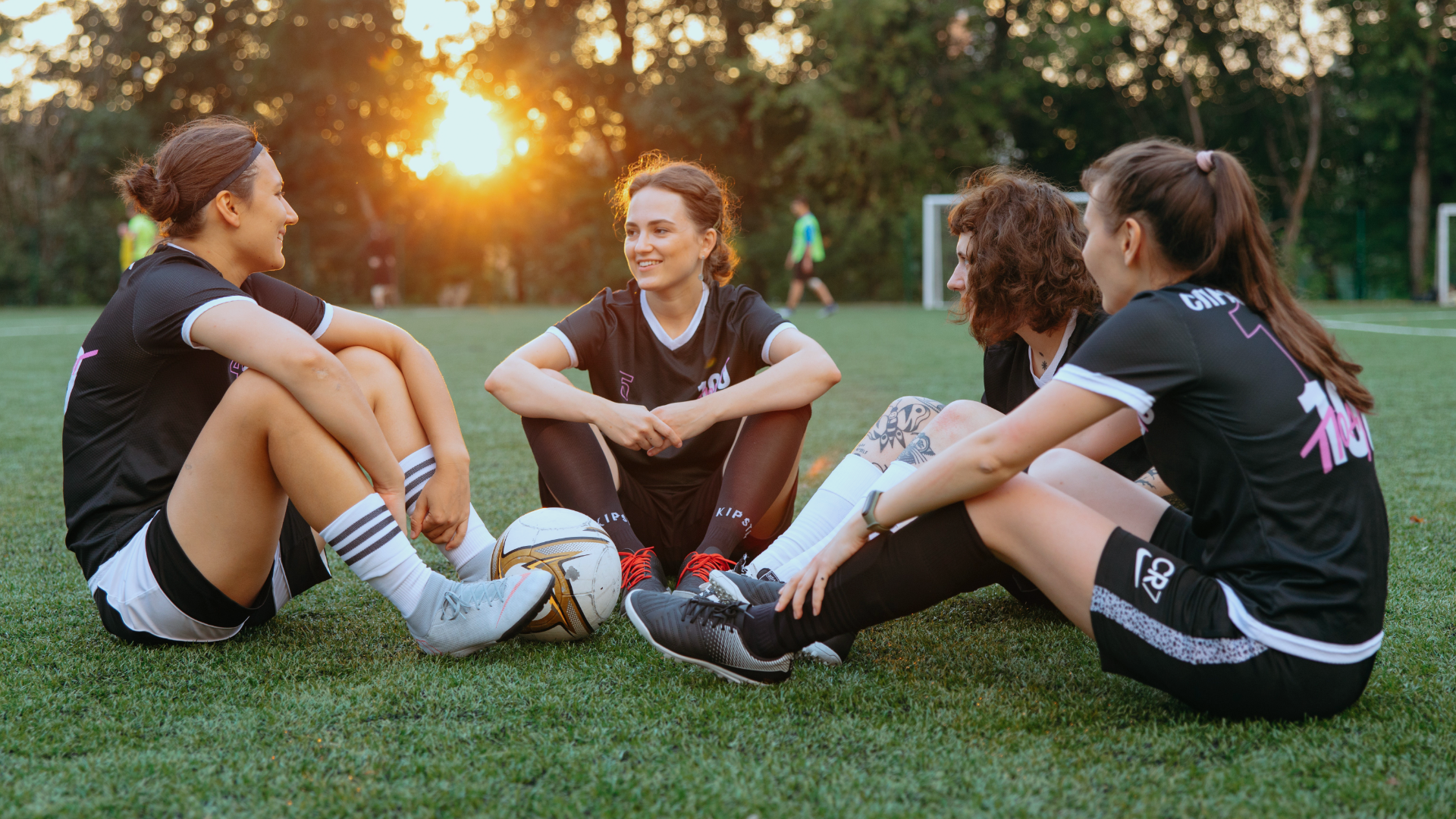 Zdjęcie przedstawia cztery zawodniczki piłki nożnej odpoczywające na murawie boiska. Dziewczyny ubrane są w piłkarskie stroje, między nimi leży piłka.