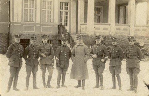 Ilustracja przedstawia grupę żołnierzy - ośmiu. Są oni ubrani w stroje wojskowe, a środkowy żołnierz również w płaszcz zimowy. Stoją oni ustawieni równo w rzędzie, pozują do fotografii, na tle budynku mieszkalnego.