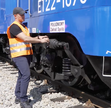 Zdjęcie przedstawia mężczyznę w kamizelce odblaskowej stojącego obok wagonu pociągu. Wskazuje on na cylinder hamulcowy.