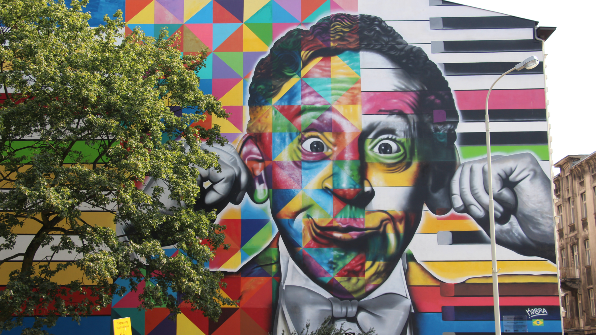 Zdjęcie przedstawia wielokolorowy mural Rubinsteina w Łodzi. Mural przedstawia twarz mężczyzny z szeroko otwartymi oczami. Mężczyzna trzyma się palcami za uszy. Po lewej stronie zdjęcia widoczne jest drzewo.