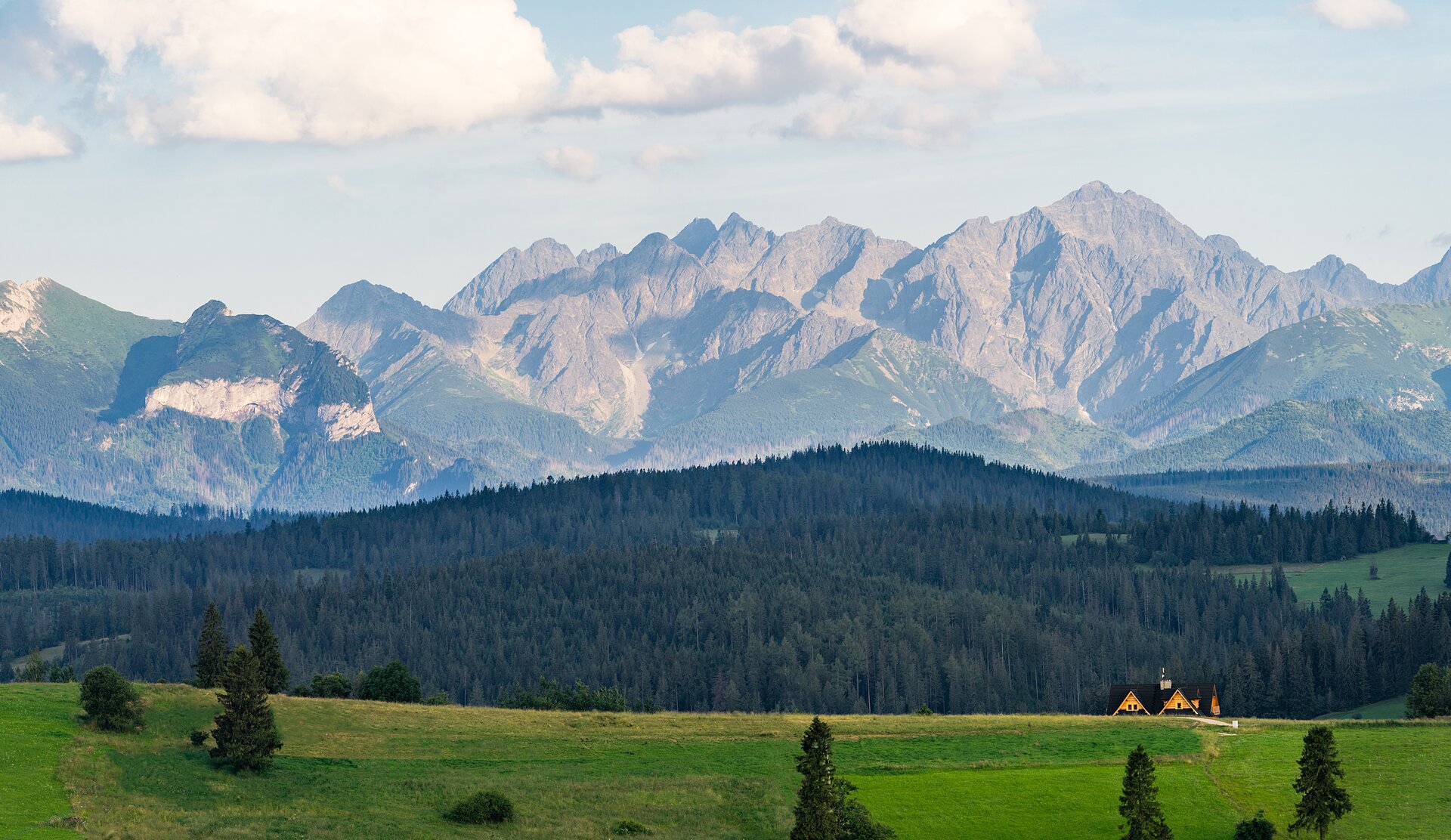 Fotografia przedstawia na pierwszym planie pole porośnięte trawą z kilkoma średniej wysokości drzewami. Dalej znajduje się wielorodzinny żółty domek ze spadzistym dachem, a za nim dolina porośnięta lasami. Za doliną znajdują się oświetlone przez słońce skalne góry.
