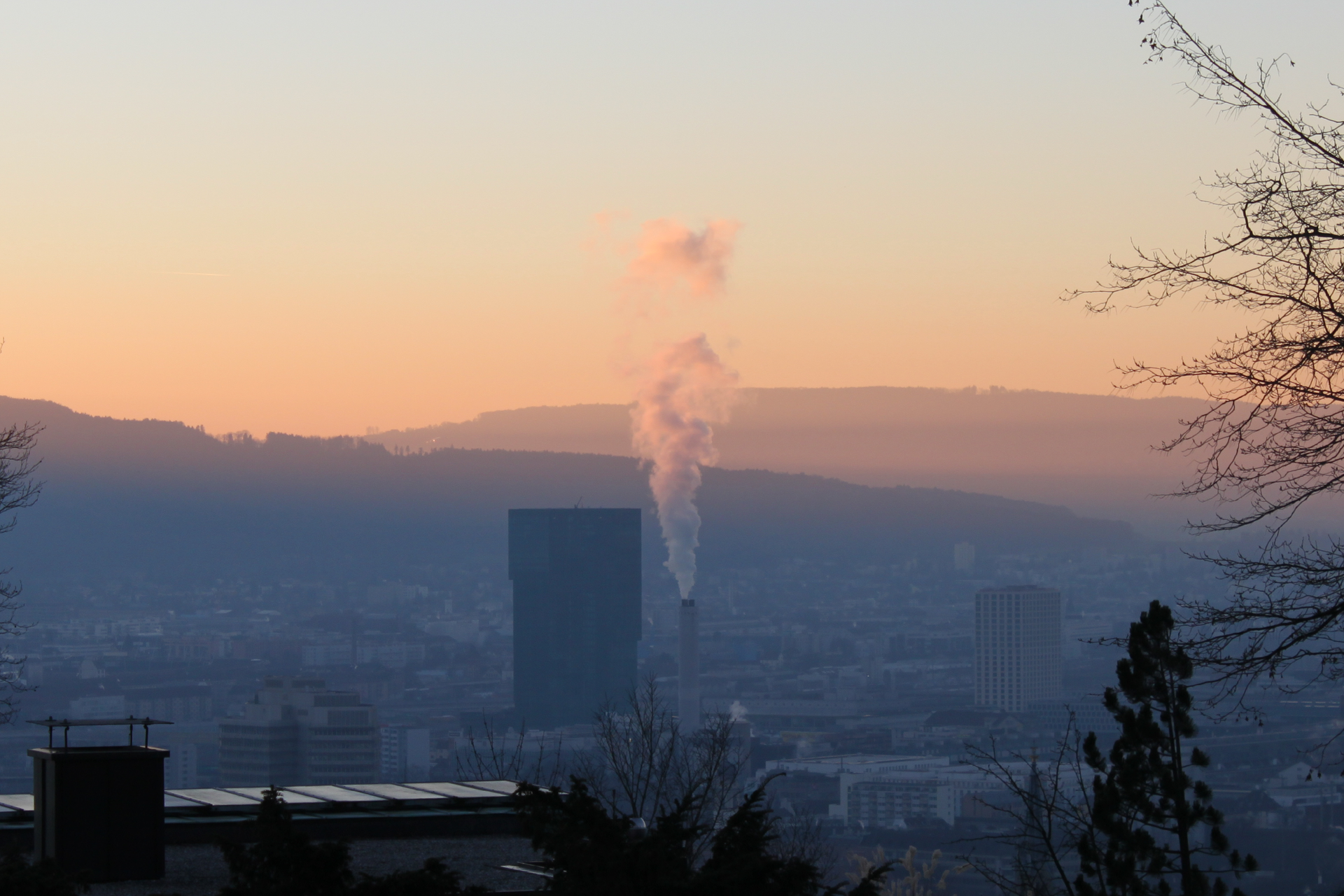Zdjęcie przedstawiające widok na miasto z oddali, w środku miasta widoczny wysoki komin, z którego unosi się prosto do góry szary dym.