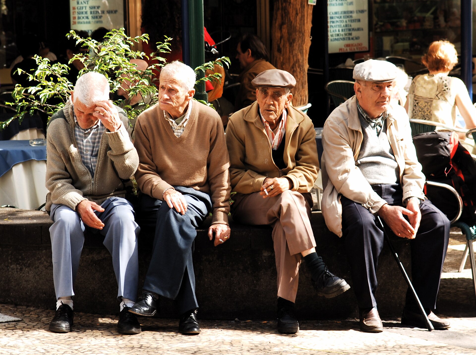 Na zdjęciu czterej mężczyźni w starszym wieku. Siwe włosy, pomarszczone twarze. Siedzą na murku przed kawiarnią. Jeden trzyma laskę.