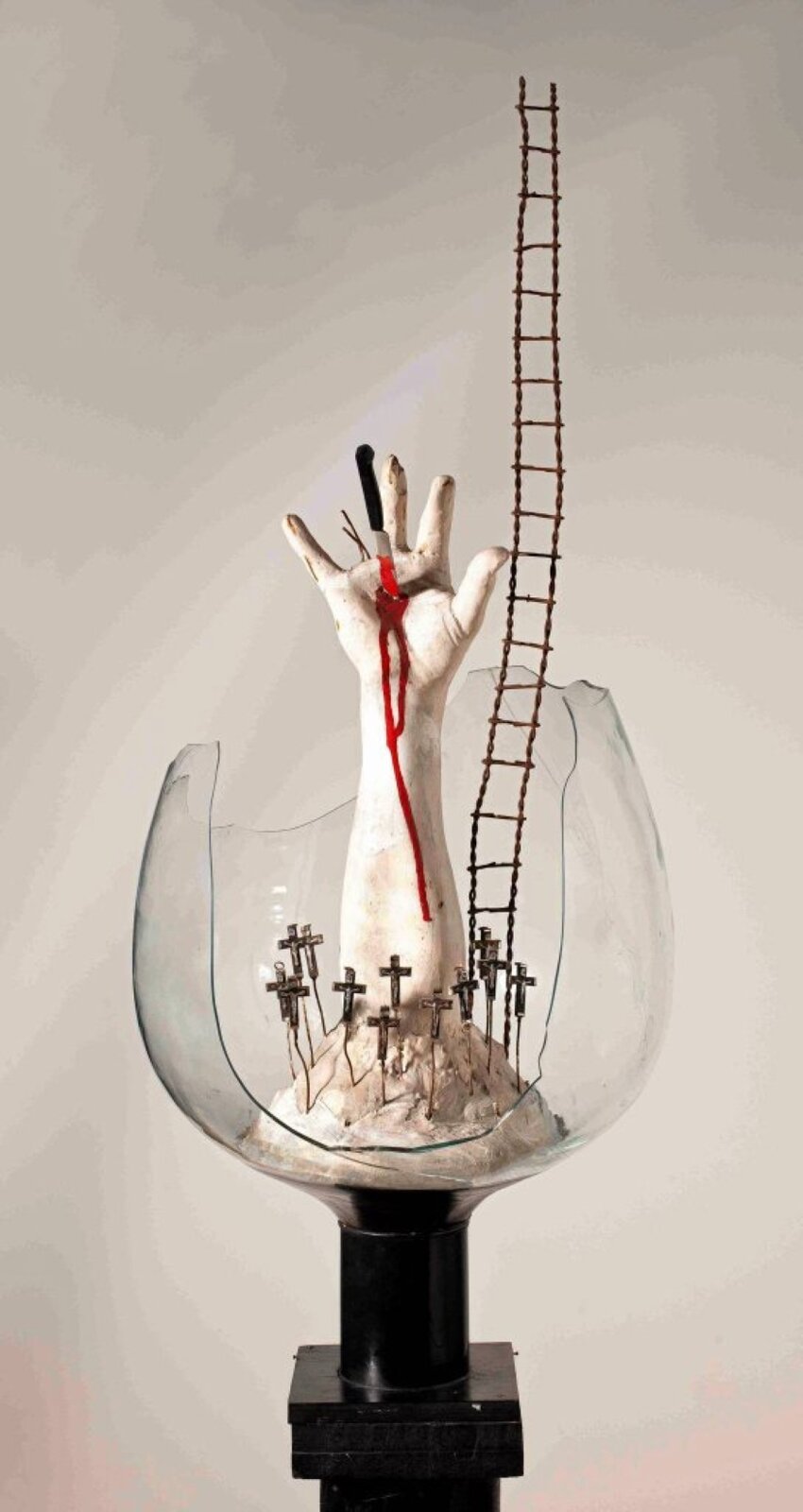 Fotografia przedstawia rzeźbę umieszczoną na czarnym podwyższeniu składającym się z elementów przypominających prostopadłościan oraz walec. Na niej umieszczony został pobity szklany kielich wewnątrz którego usypano jasne wzgórze, na którym rozmieszczono krzyże stojące na drucianych nóżkach. Otaczają one wyłaniającą się ze szczytu góry jasną, ludzką rękę, której palec serdeczny zastąpiono dwoma krótkimi kabelkami. Pośrodku otwartej dłoni umieszczono wbity nóż o czarnej rączce oraz srebrnym, zakrwawionym ostrzu. Krew cieknie z rany, sięgając prawie końca przedramienia. Z prawej strony dłoni stoi wysoka pokrzywiona drabina. Tło stanowi rozświetlona biała ściana.