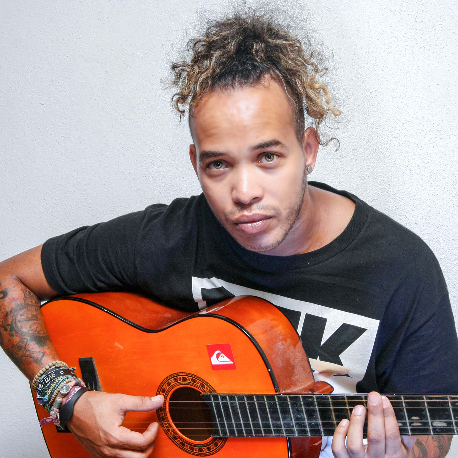 Zdjęcie przedstawia nastolatka z upiętymi w kucyka włosami. Chłopak ma wytatuowane ręce, trzyma gitarę, a jego palce muskają struny. 