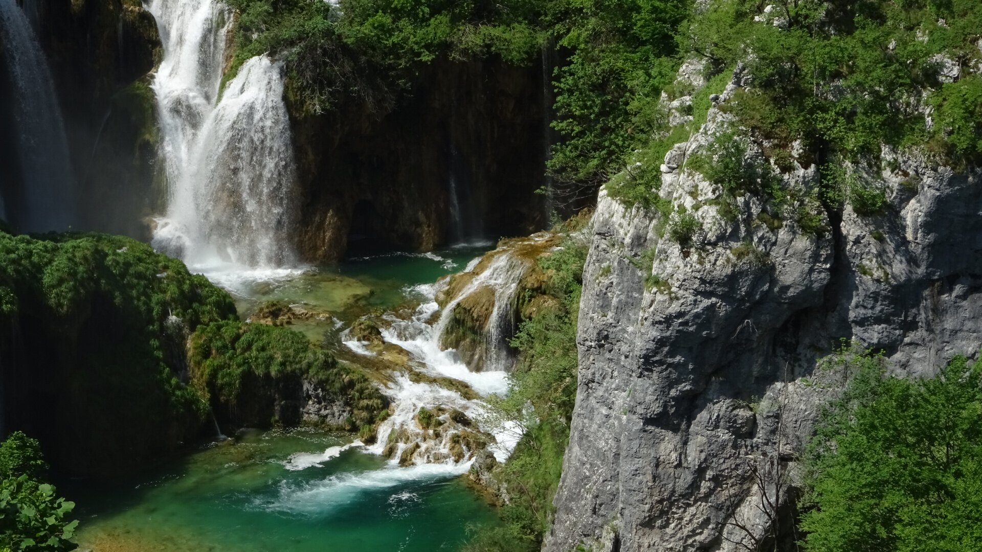 Po prawej stronie zdjęcia widać górę złożoną z szarych pionowych skał porośniętą zieloną roślinnością. Po jej lewej stronie znajdują się półki skalne, po których spływa woda, tworząc niewielkie kaskady. Poniżej jest nieduże jeziorko, a powyżej większe jezioro i wysokie ściany skalne, z których dwoma strumieniami spływa woda, tworząc wodospady. Po lewej widać niewielkie skały porośnięte zieloną roślinnością.
