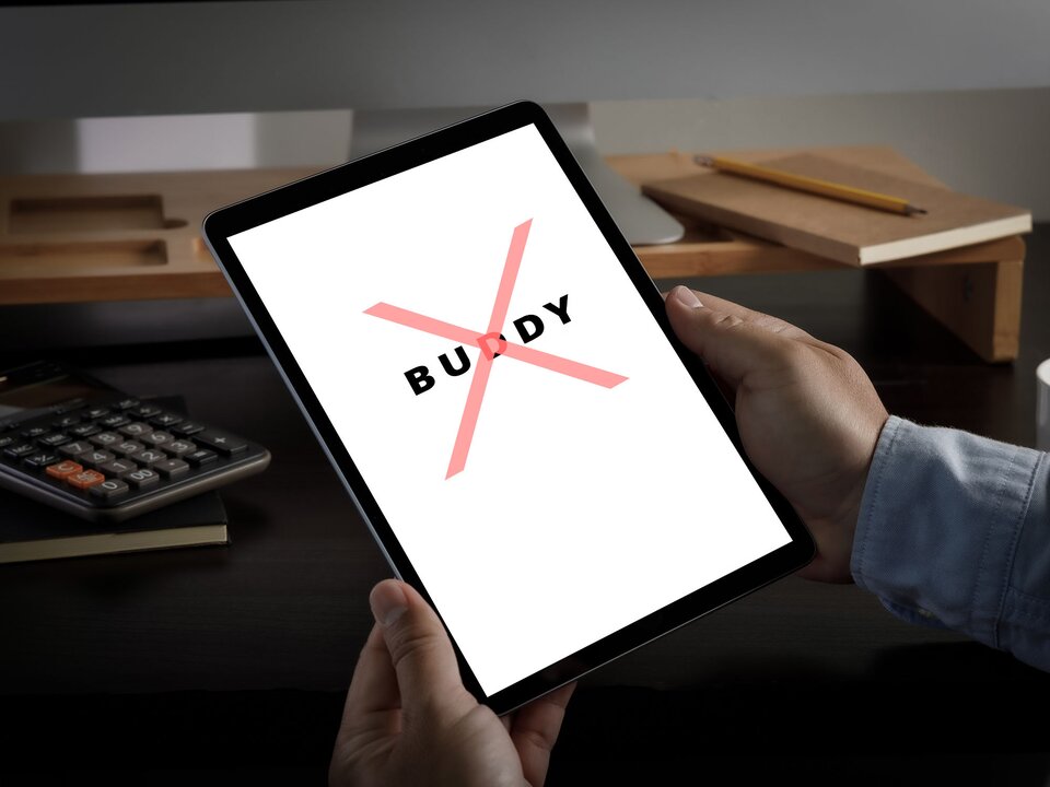 Zdjęcie przedstawia tablet, który trzymany jest w dłoniach. Na ekranie tabletu widoczny jest napis z czarnych liter w języku angielskim: BUDDY przekreślony dużym, czerwonym znakiem X. W tle widoczny fragment biurka z kalkulatorem i notesem, na którym leży ołówek.