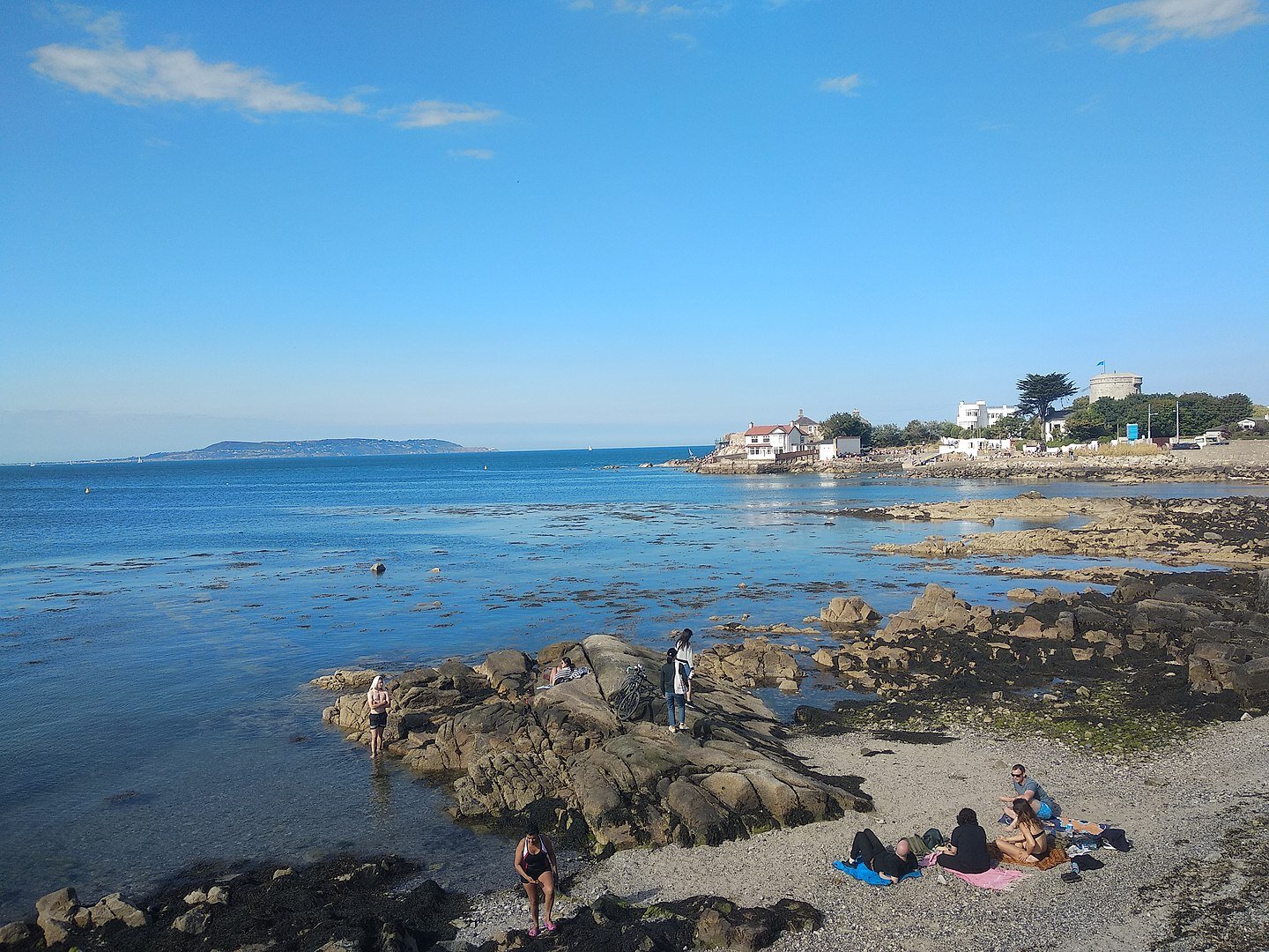 Fotografia przedstawiająca nadmorską, kamienistą plażę. Na pierwszym planie wypoczywający ludzie, ubrani w kostiumy kąpielowe. Na drugim planie, po prawej stronie niskie zabudowania. W tle zarys wyspy.