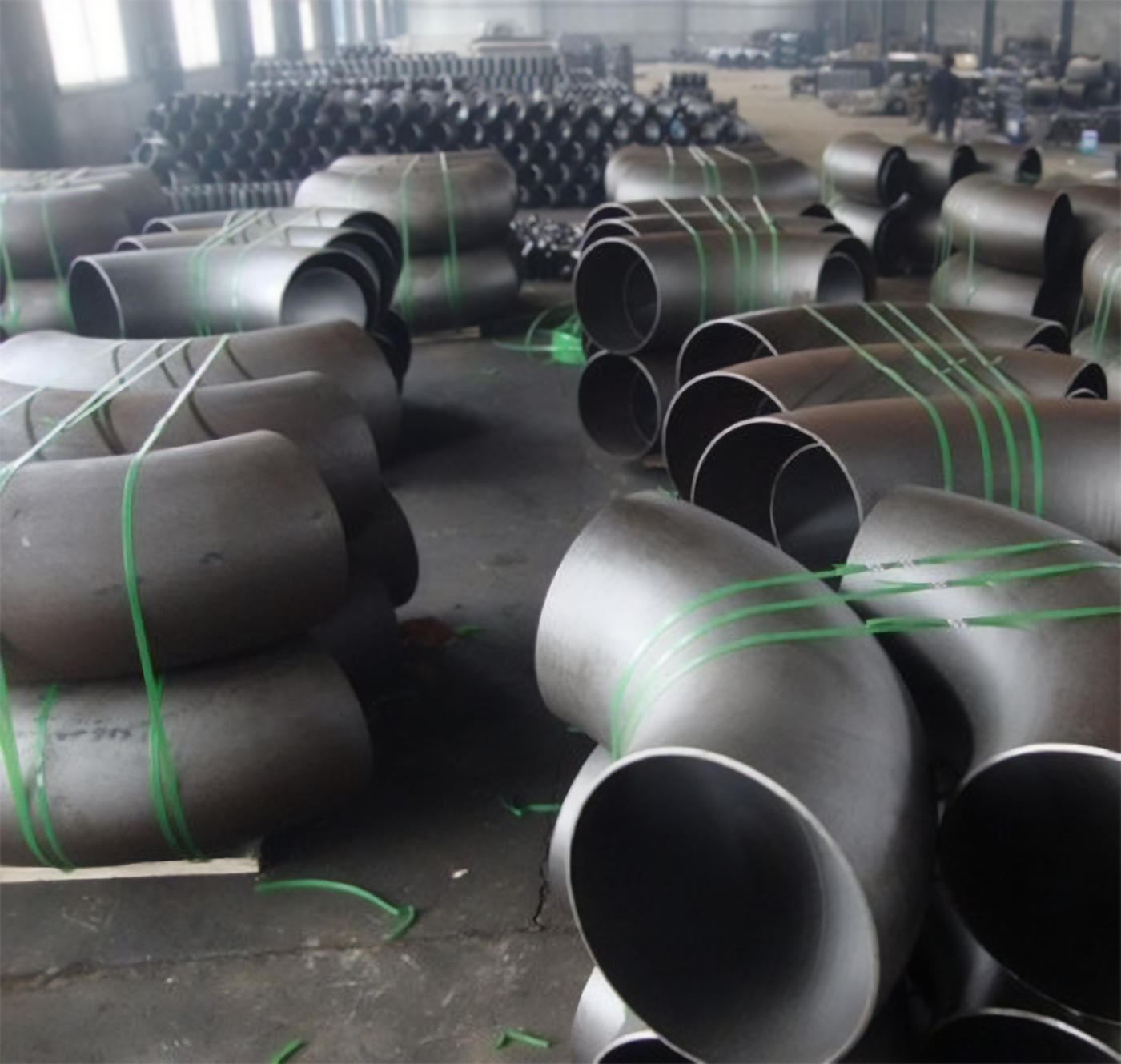 Zdjęcie przedstawia przygotowanie kolan metalowych do transportu. Widać duże kolana, które mają postać lekko zgiętych rur. Są one ułożone na palecie, przywiązane są do niej pasami.