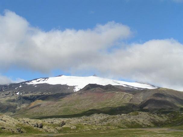 Fotografia wykonana u stóp dużego, płaskiego wzniesienia. Dolne partie pokryte trawą, górne tworzą gołe ciemnobrązowe skały, szczyty ośnieżone.