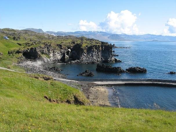 Na zdjęciu pionowy, wysoki klif o ciemnoszarej barwie, u góry lekko pagórkowaty teren pokryty trawą. Przed lądem spokojne morze.