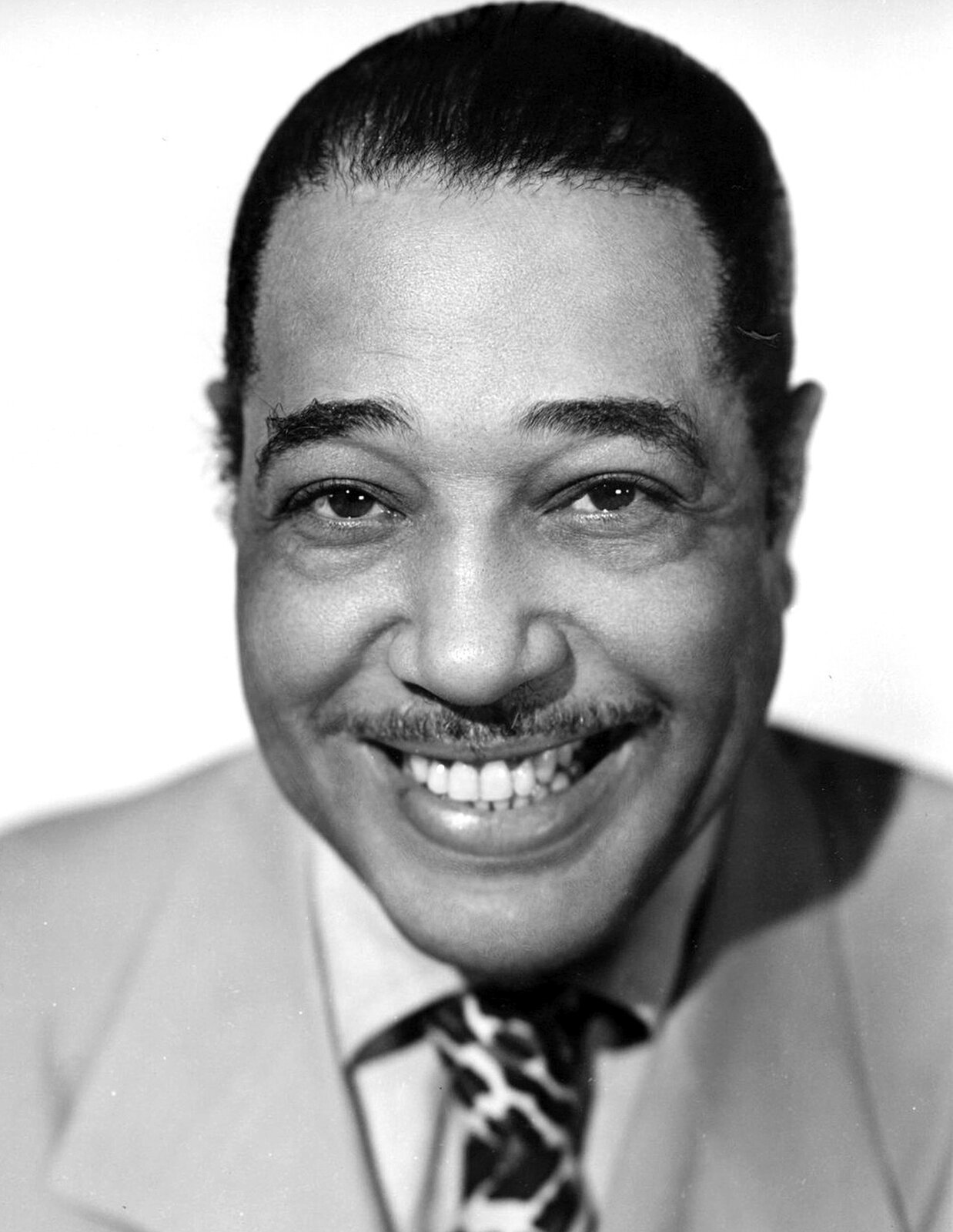 Na zdjęciu Duke Ellington. Czarnoskóry mężczyzna szeroko się uśmiecha wprost do aparatu. Muzyk ubrany jest w koszulę, łaciaty krawat i jasną marynarkę.
