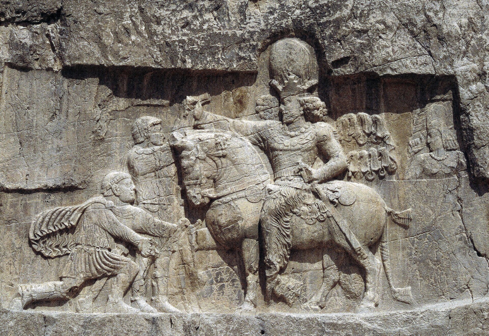 Fotografia przedstawia relief ukazujący trzy postacie, w tym jedną konno, w zbroi oraz z mieczem przy boku. Głowa jeźdźca ozdobiona jest wysokim nakryciem głowy, spod którego wystają kręcone włosy. Prawą rękę mężczyzna wyciąga do dwóch pozostałych postaci – jednego stojącego przed nim w hełmie oraz drugiego klęczącego mężczyznę z workiem niesionym na plecach. Tło stanowi zadrapana szara ściana skalna.