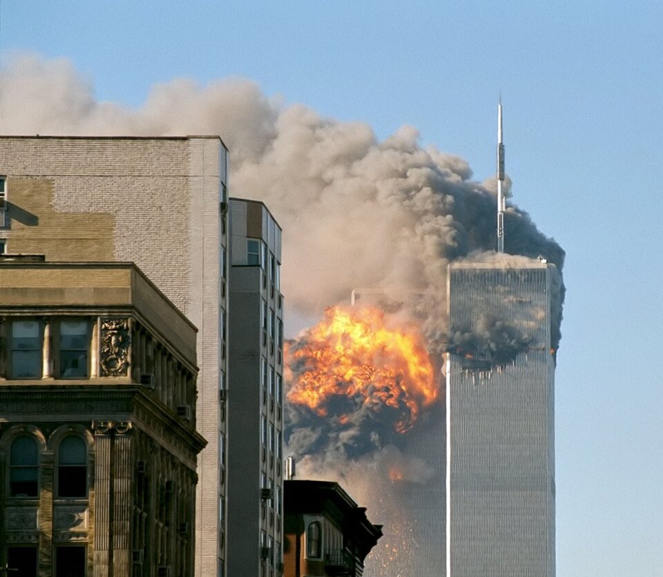 kliknij aby powiększyć Zdjęcie przedstawia atak terrorystyczny na dwie wieże budynku World Trade Center w Nowym Jorku 11 września 2001 roku. Widoczne są płonące wieżowce i wydobywające się ogromne kłęby dymu po uderzeniu samolotów. Z lewej strony stoją gmachy innych wysokich budynków. W tle zdjęcia widać niebo.
