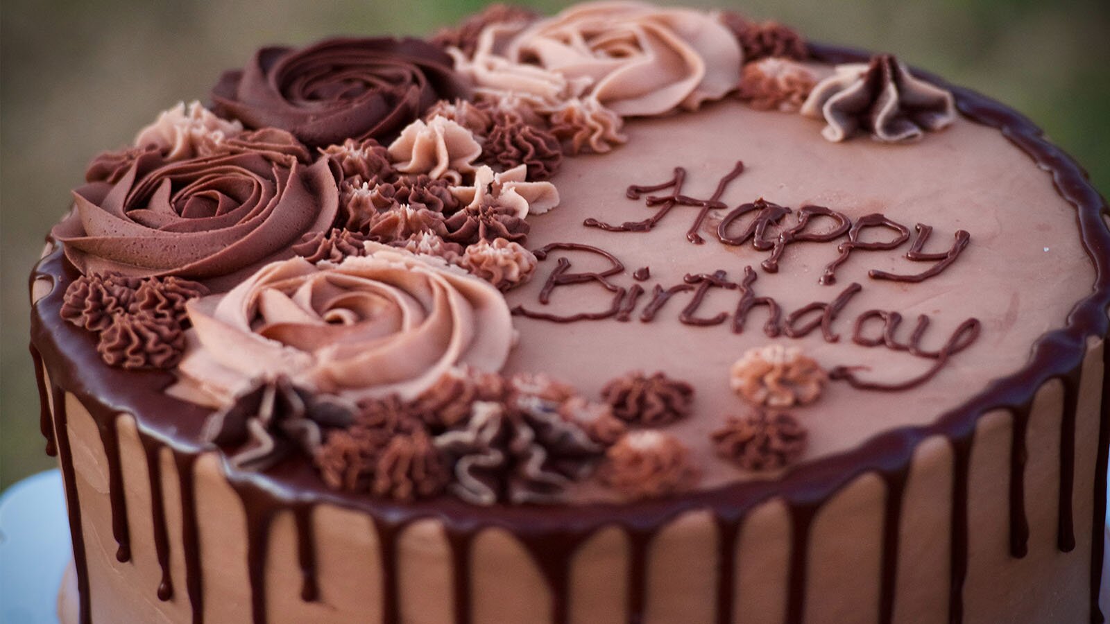 Zdjęcie przedstawia tort czekoladowy, na którym widoczny jest napis w języku angielskim: HAPPY BIRTHDAY. Na lewo od napisu, a także u góry i u dołu, widoczne są kwiaty wykonane z czekolady w kolorze od różowego do ciemnobrązowego.