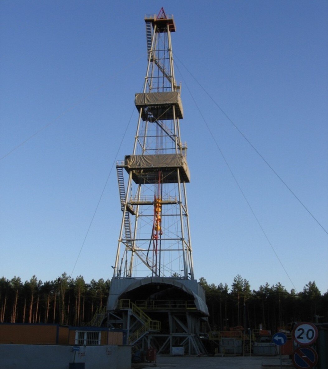 Na zdjęciu widać wieżę wiertniczą. Widoczne wieża jest konstrukcją kratową o kształcie ostrosłupa ściętego. Wieża zbudowana jest z trzech galerii. Do każdej galerii prowadzą schody. Galerie po obwodzie są otoczone szarym płótnem. U podstawy wieży widać betonowy plac wiertniczy.W tle zdjęcia znajduje się las.