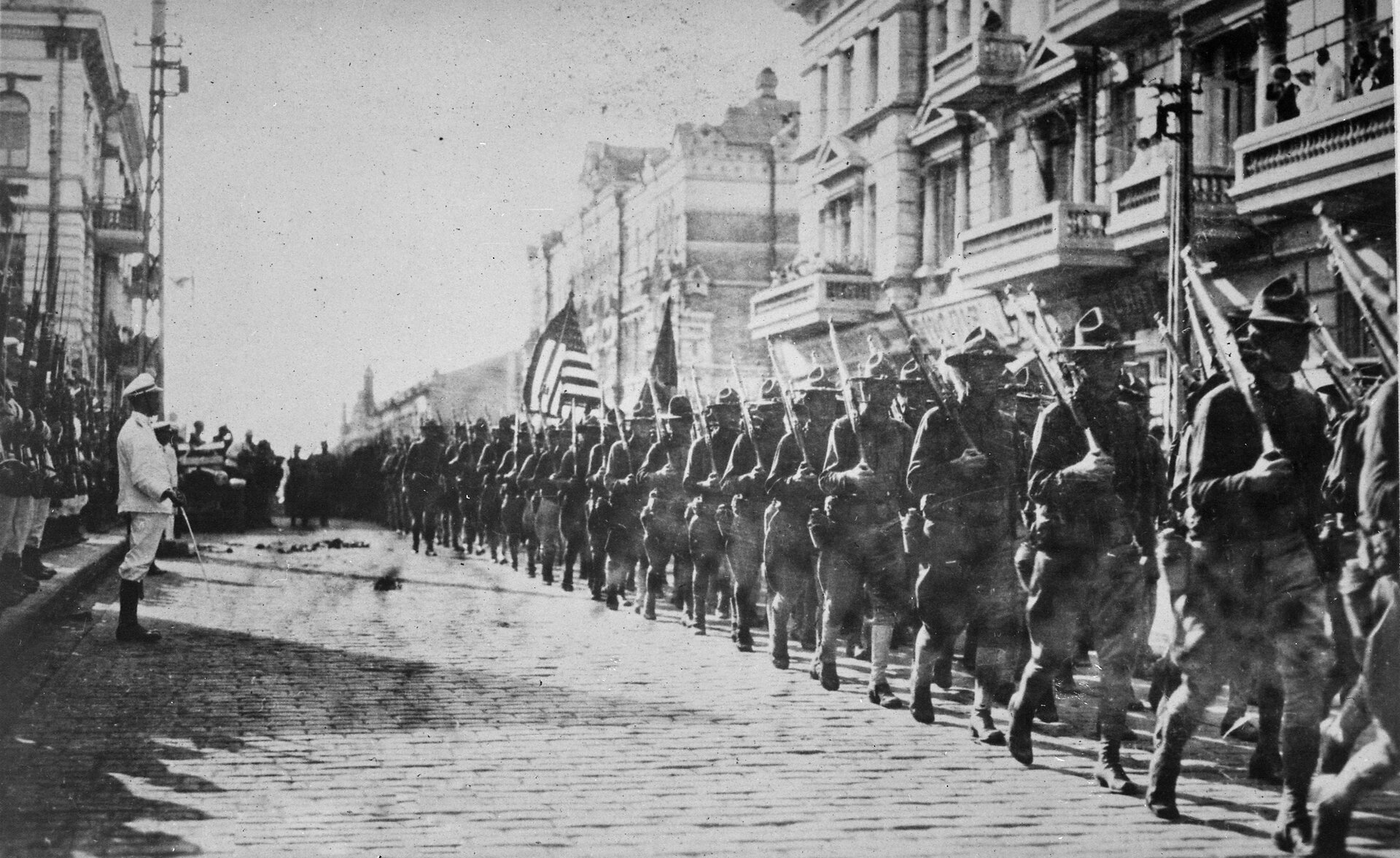 Na zdjęciu przemarsz oddziału wojskowego armii amerykańskiej. Przemarsz odbywa się ulicą miasta, po obu stronach ulicy znajdują się kilkupiętrowe budynki. Żołnierze po prawej stronie zdjęcia ubrani są w mundury oraz kapelusze, trzymają na ramionach karabiny. Wśród maszerujących żołnierzy znajduje się chorąży trzymający flagę USA. Defiladę ogląda wyższy stopniem oficer (widoczny po lewej stronie zdjęcia),  za którym stoi w postawie na baczność oddział żołnierzy, którzy prezentują broń. W tle znajduje się otwarty samochód osobowy, przy którym stoi kilkuosobowa grupka żołnierzy.  